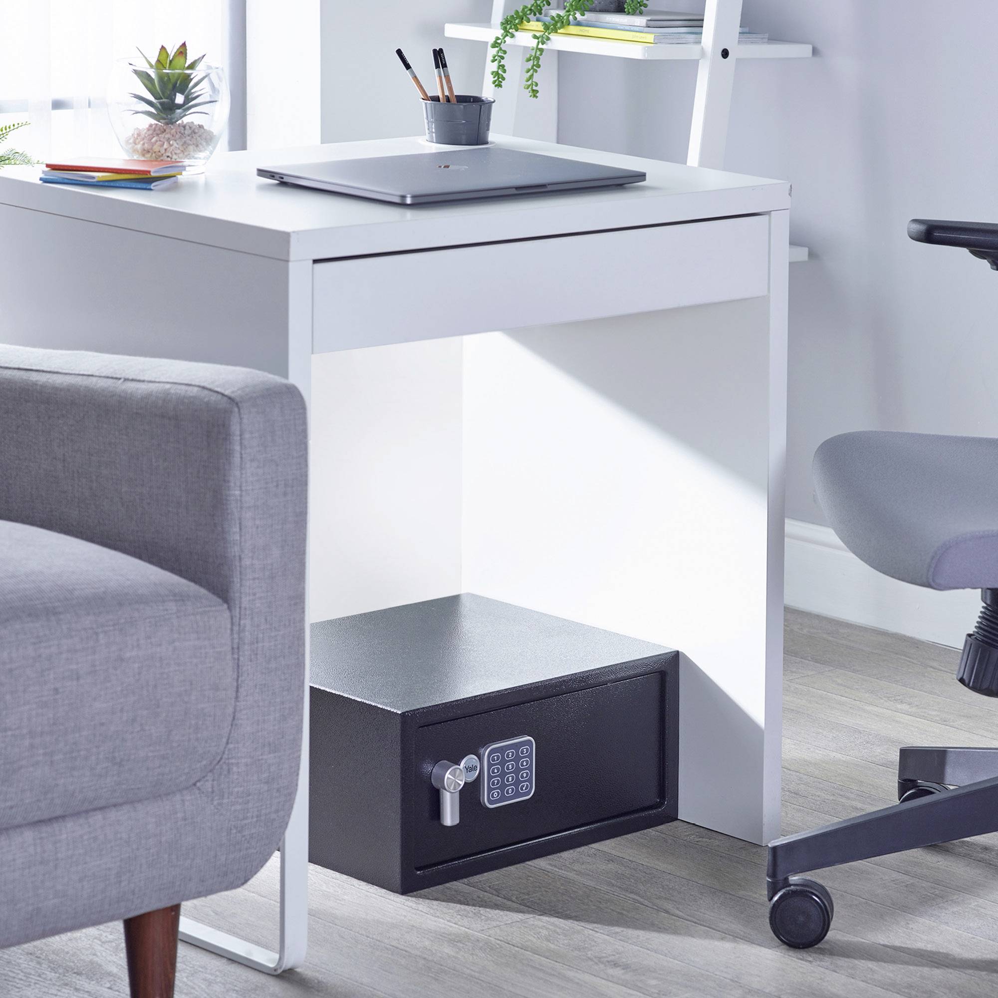 Un bureau blanc avec un ordinateur portable et des plantes est installé dans un bureau moderne. Un coffre-fort se trouve sous le bureau.