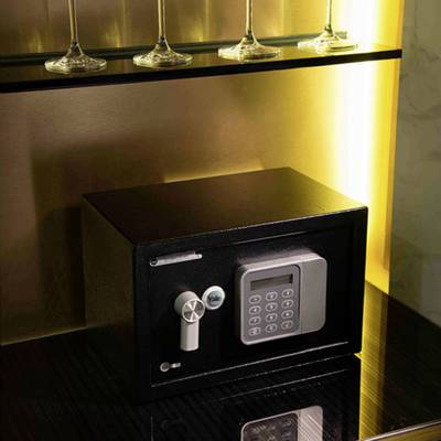 Un coffre-fort à serrure électronique est posé sur une étagère en bois, sous une rangée de verres à vin, discrètement éclairé.