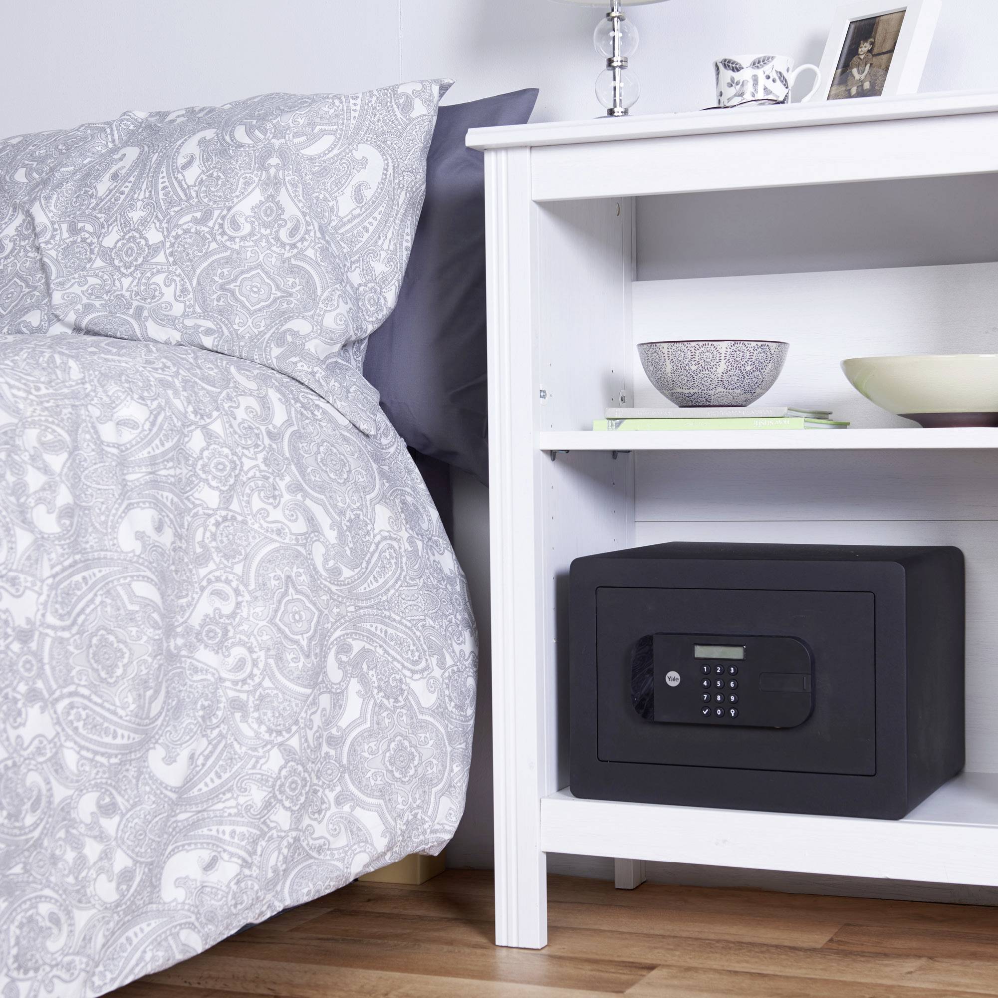 Une chambre avec un lit à gauche et une table de nuit blanche à droite. Sur la table se trouve un coffre-fort dans une étagère.