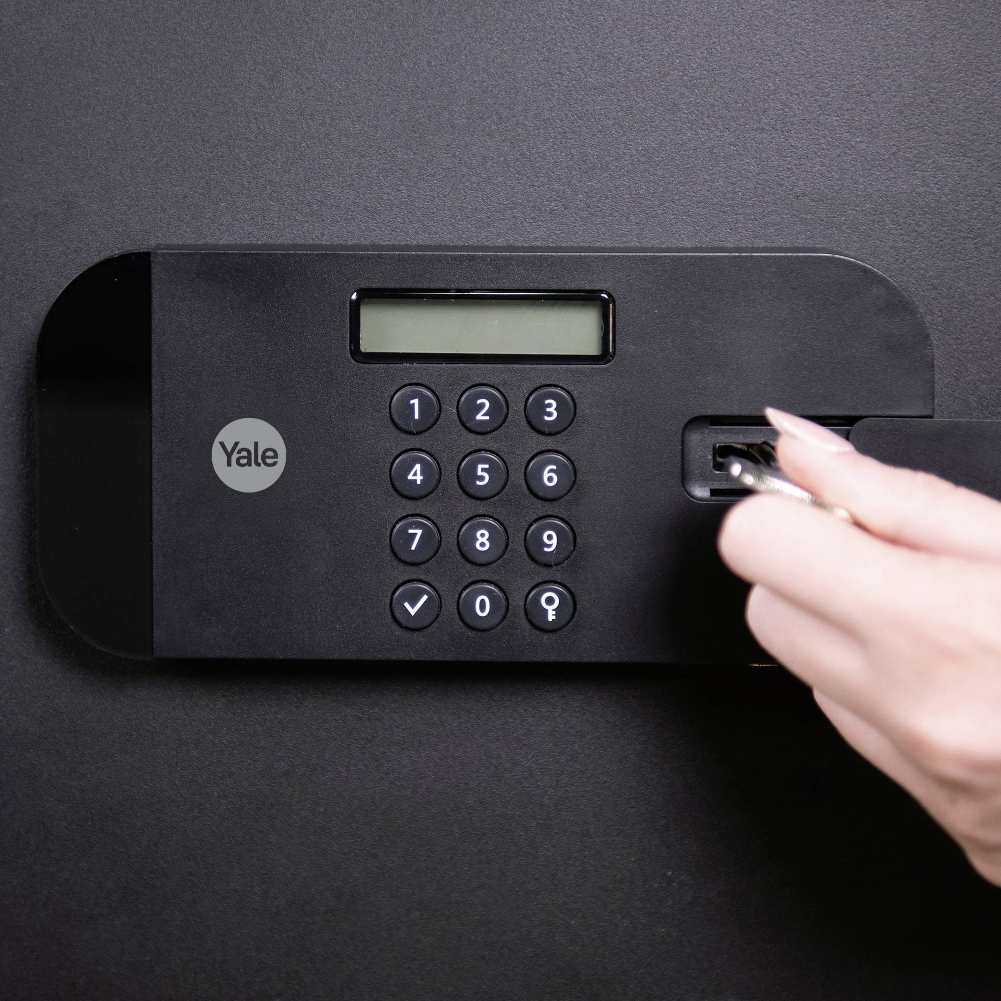 Une main entre un code sur un pavé numérique d'un verrou électronique sombre fixé au mur.