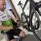 Une personne en tenue de cyclisme installe un cadenas noir sur un vélo de course. Elle est assise par terre à côté d'un bâtiment.