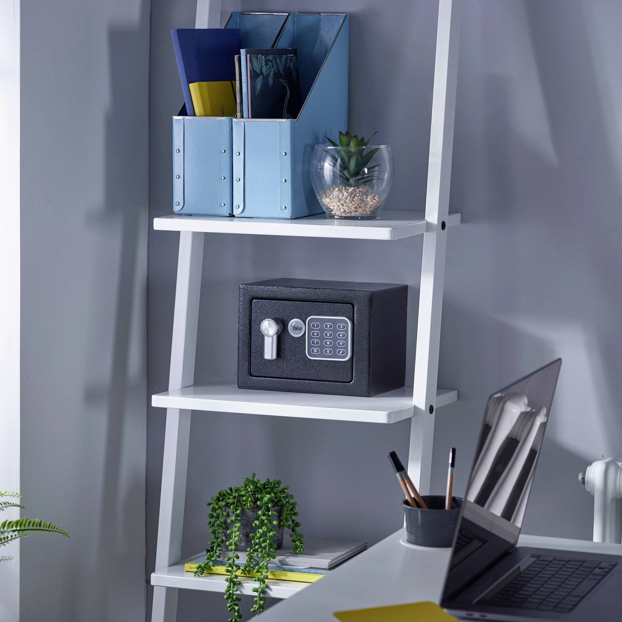 Une étagère blanche avec un petit coffre-fort, des classeurs de bureau et des plantes posés dessus dans un bureau moderne. Une table avec des stylos est placée devant.