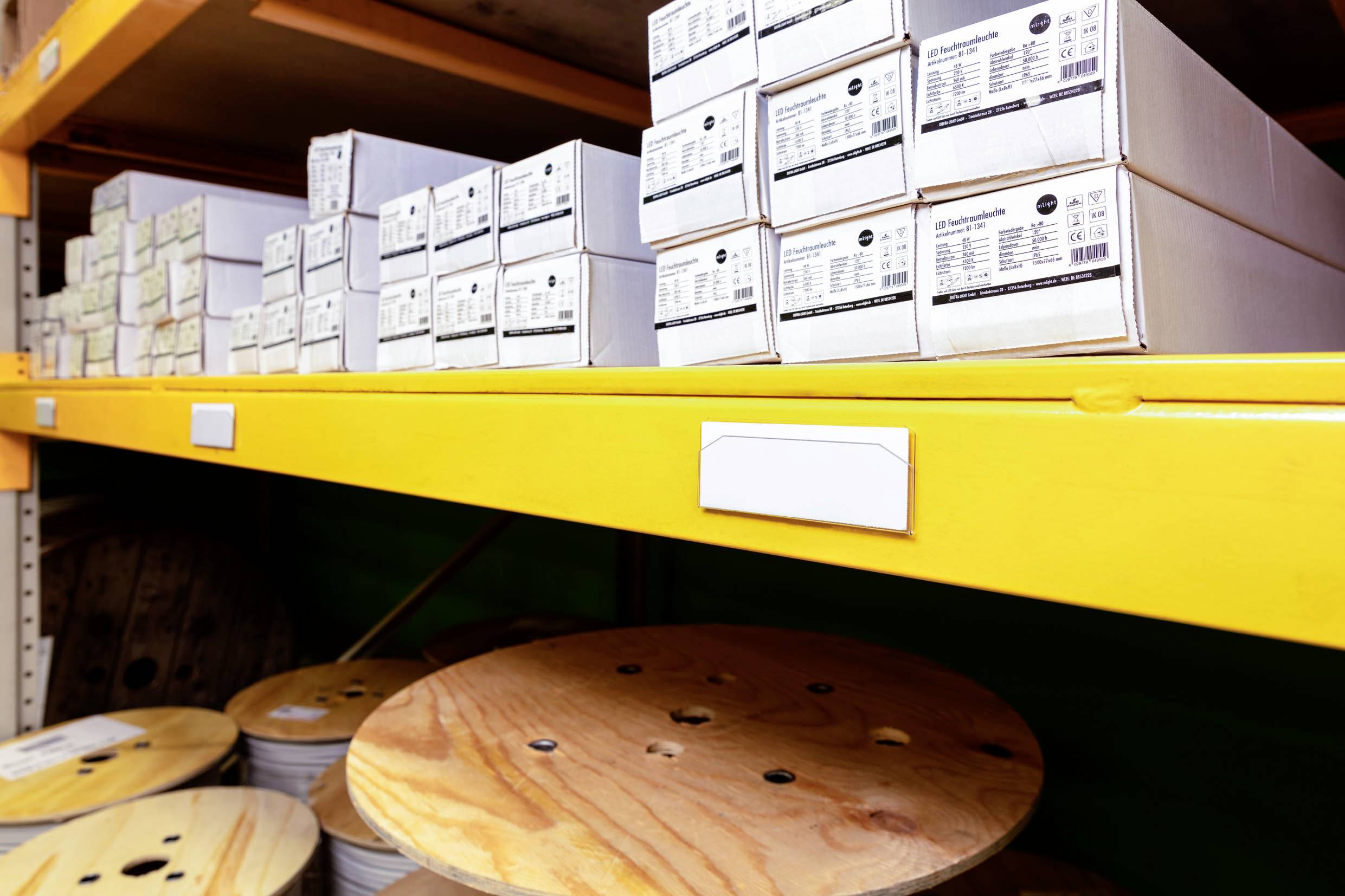 Étagères jaunes avec des boîtes blanches empilées et des bobines en bois dans un entrepôt. Les boîtes sont étiquetées.
