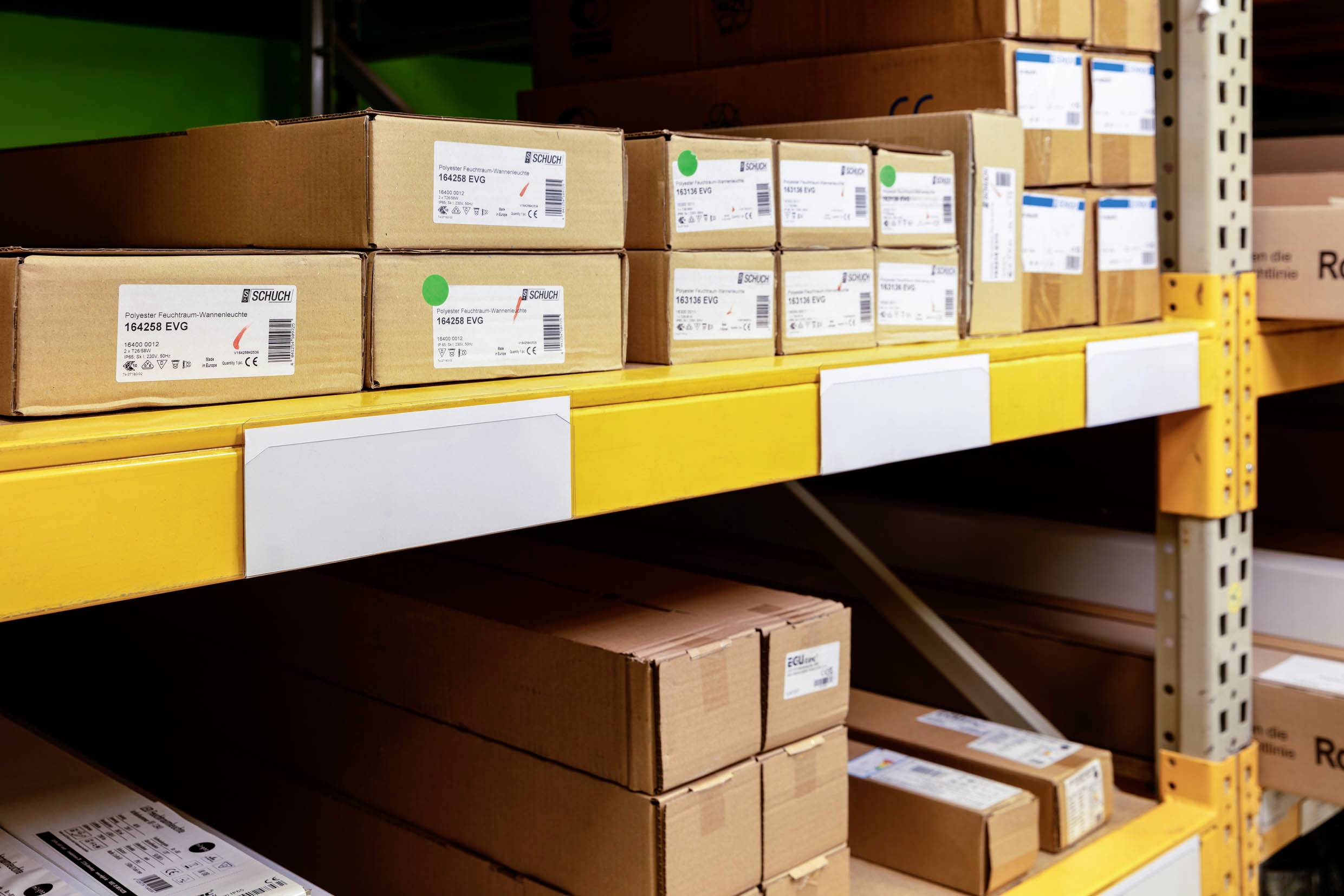 Étagères dans un entrepôt avec des boîtes en carton brun empilées. Des étiquettes sont visibles, portant des spécifications techniques.