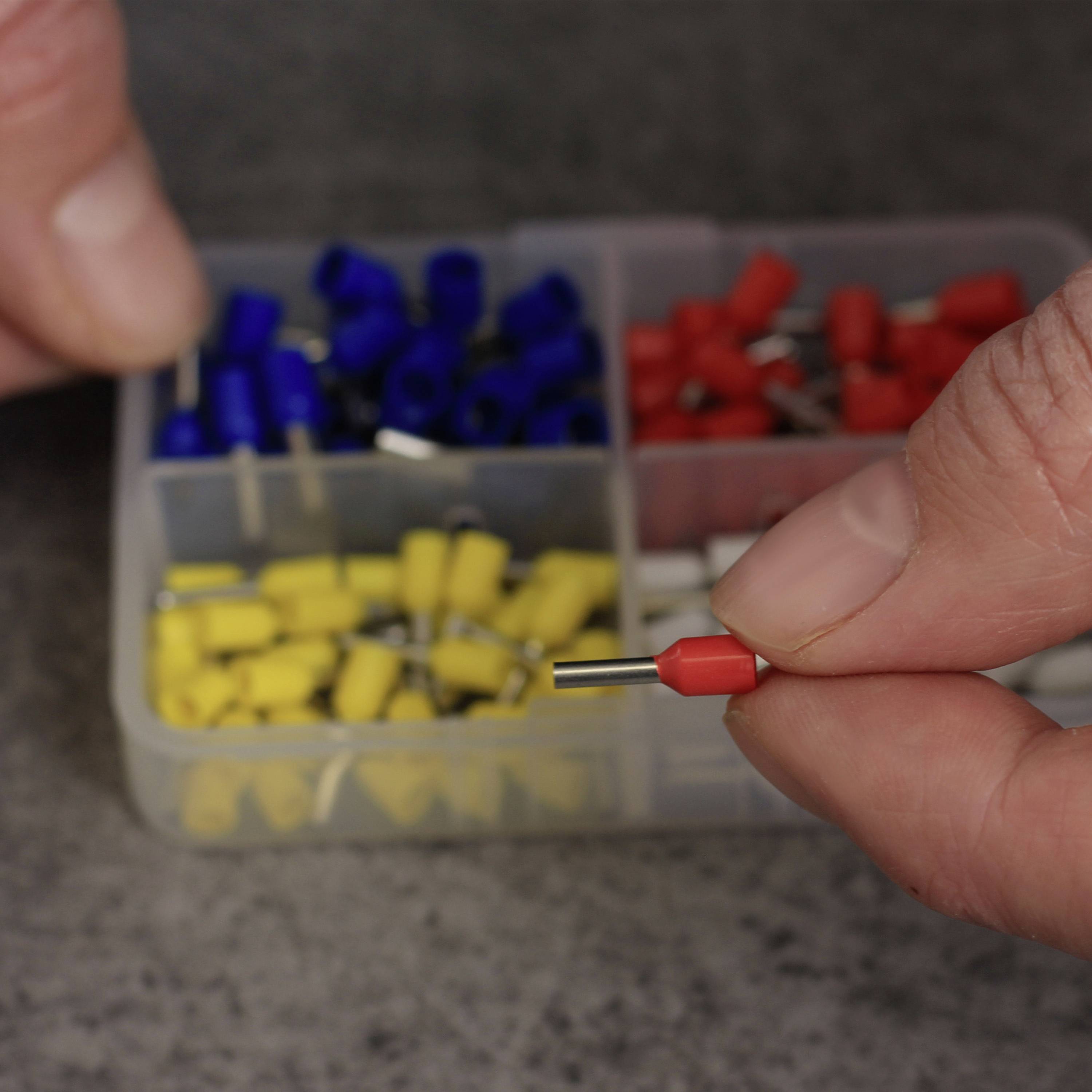 Deux mains, l'une tenant une fiche de câble jaune, tandis que l'autre pointe une fiche rouge dans une boîte en plastique remplie de fiches de différentes couleurs.