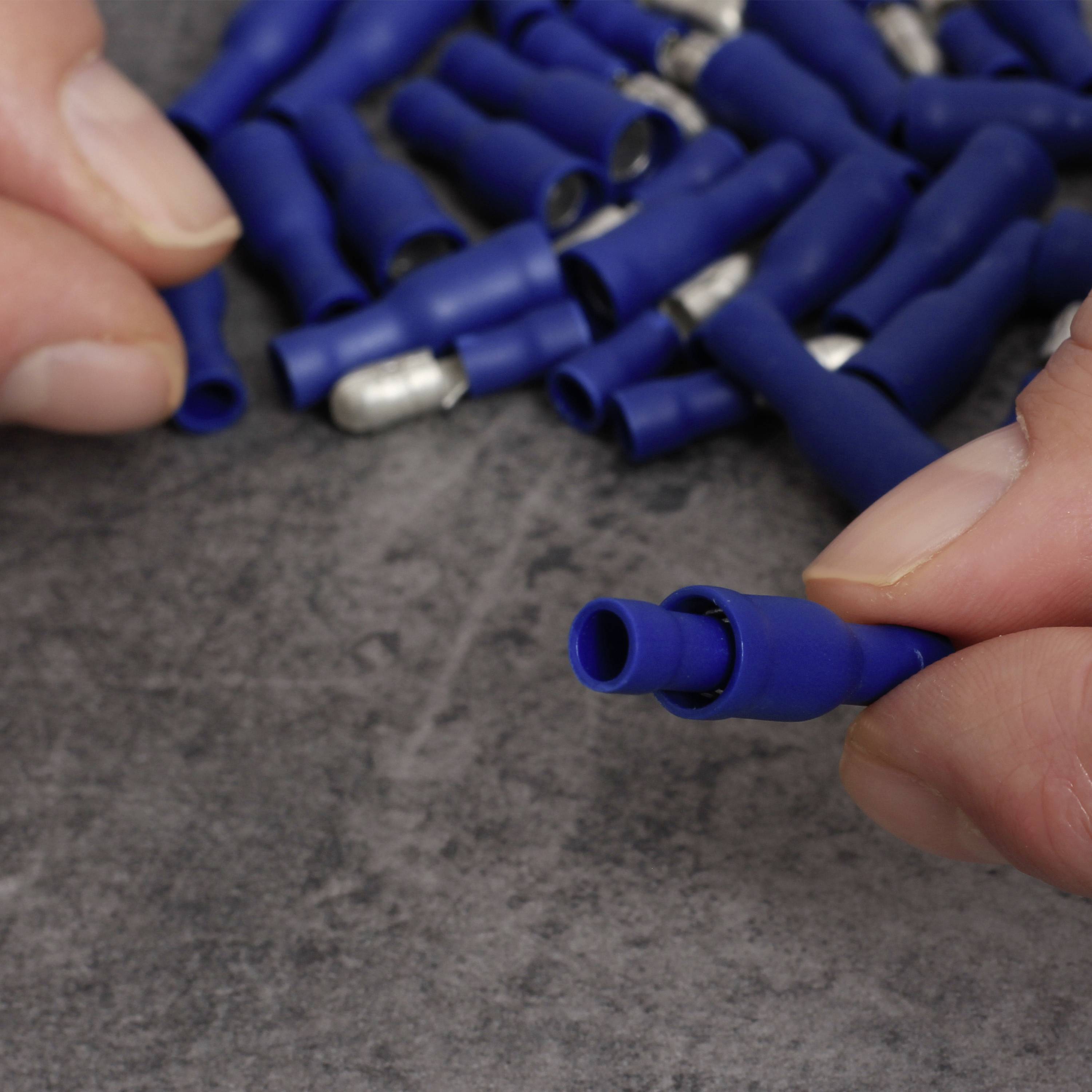 Des mains tiennent une cosse de câble bleue, entourée d'autres cosses sur une table. Cela sert à connecter des câbles électriques.