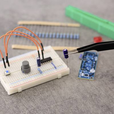 Une plaquette de circuit imprimé avec des câbles, des résistances et une puce de microcontrôleur. Une pince maintient un condensateur. Des composants électroniques sont visibles en arrière-plan.
