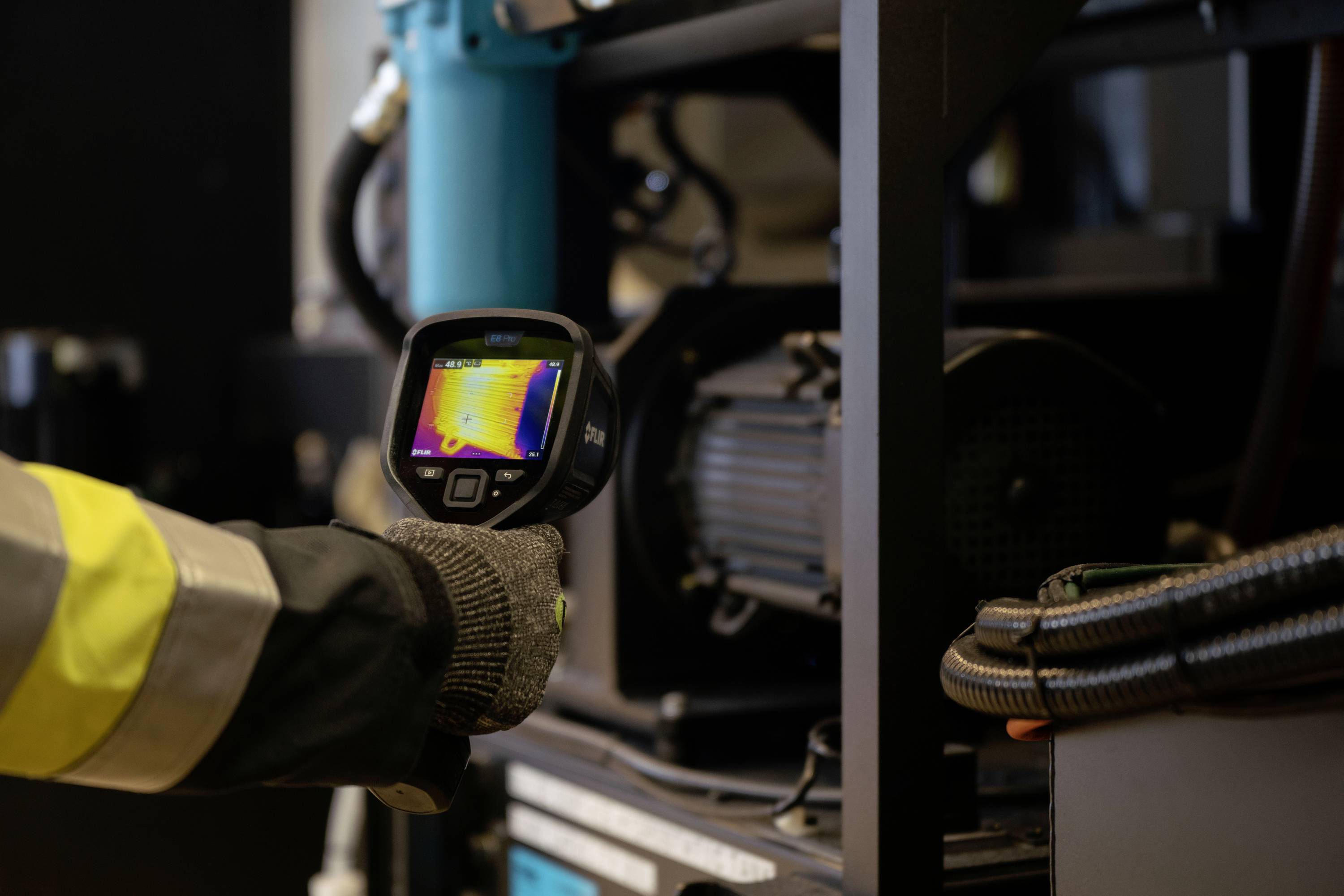 Une personne tient une caméra thermique à la main, dirigée vers une machine. L'écran affiche une image thermique colorée.