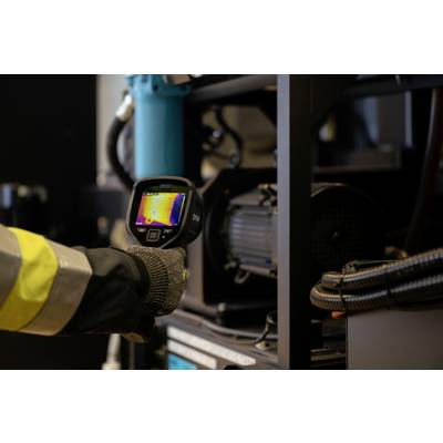 Une personne tient une caméra thermique à la main, dirigée vers une machine. L'écran affiche une image thermique colorée.