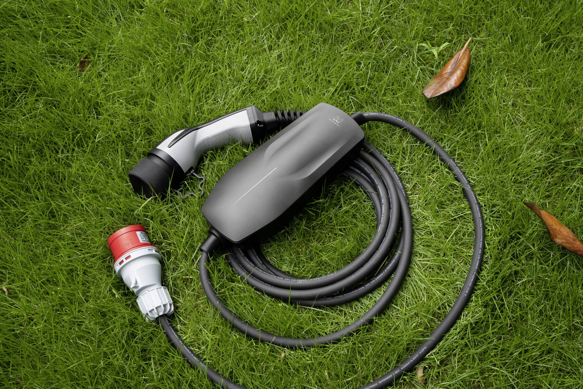 Un câble de charge noir pour un véhicule électrique est posé sur de l'herbe verte. Il possède une poignée en plastique et une prise.