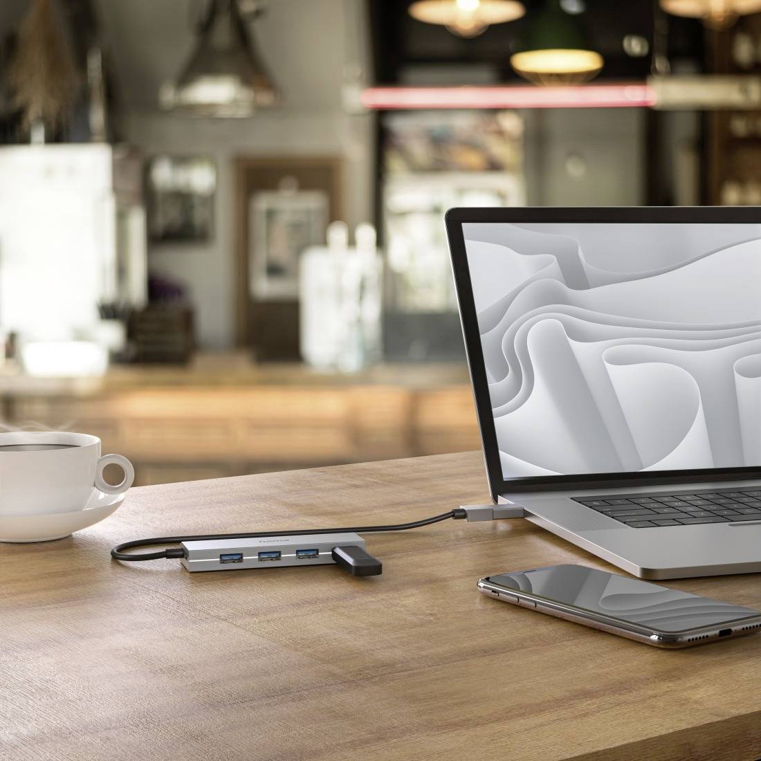Un ordinateur portable posé sur une table de café, connecté à un hub USB, à côté d'un smartphone et d'une tasse de café. Arrière-plan flou dans l'ambiance d'un café.