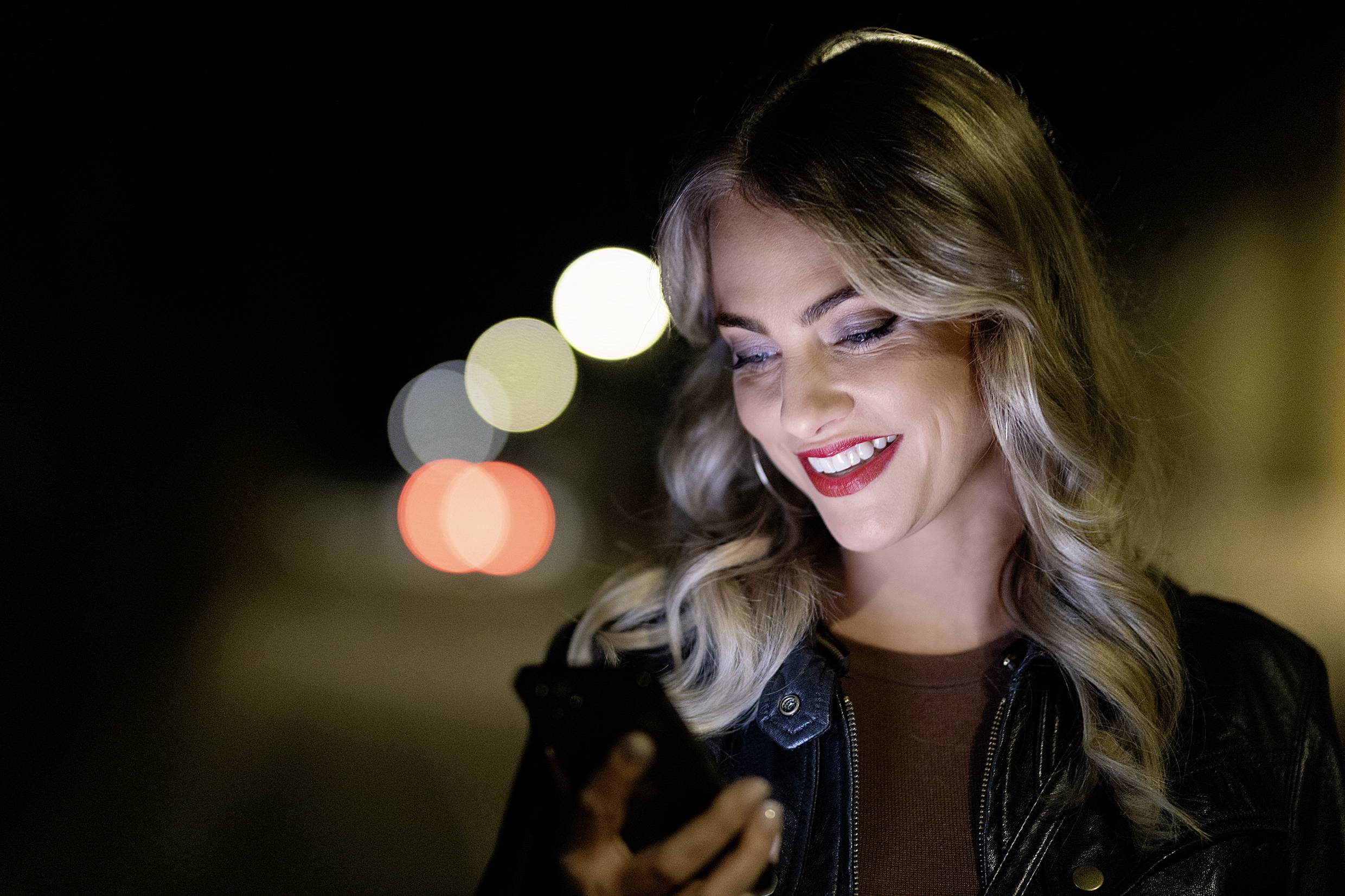 Une femme souriante aux cheveux blonds regarde son smartphone alors qu'elle se trouve dehors la nuit. Des lumières brillent en arrière-plan.