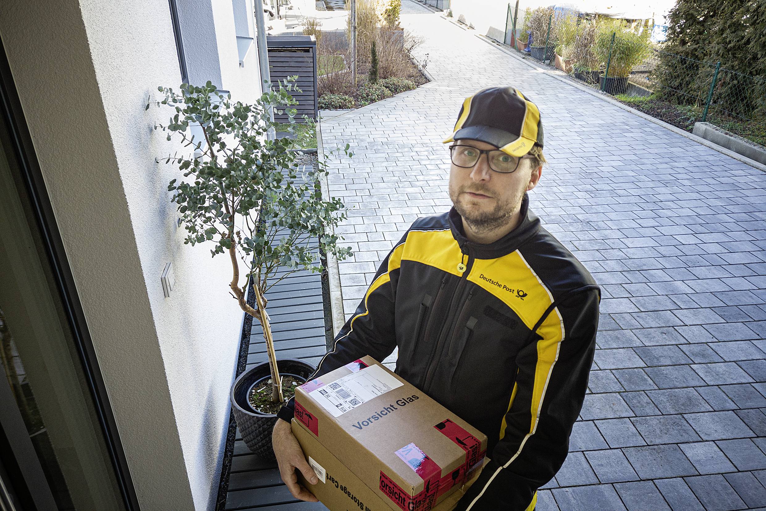 Un livreur en uniforme jaune et noir tient deux colis. À l'arrière-plan, on aperçoit un trottoir pavé et un petit arbre.