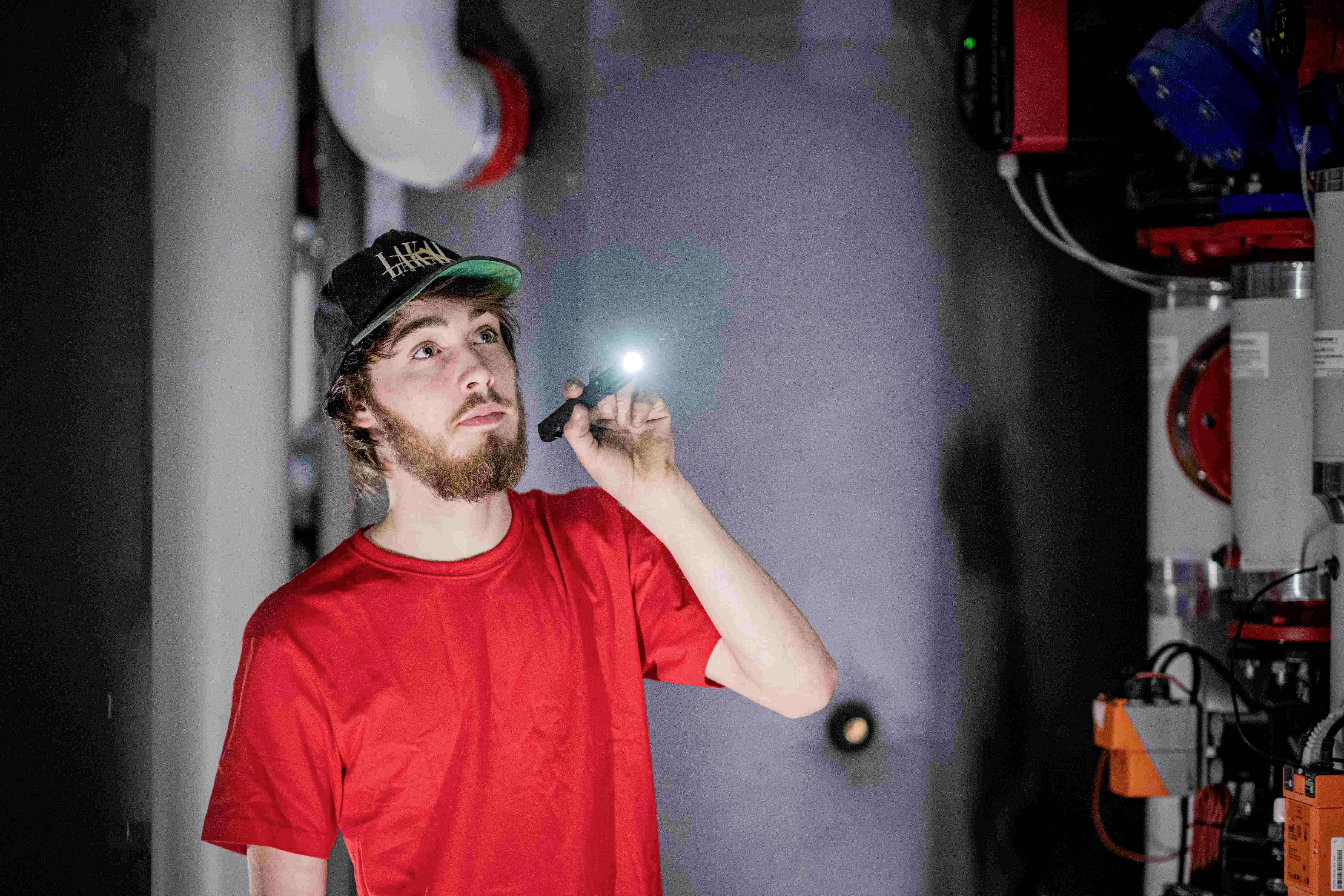 Un homme portant un t-shirt rouge éclaire la pièce avec une lampe de poche dans une salle sombre où des appareils techniques sont installés sur les murs.