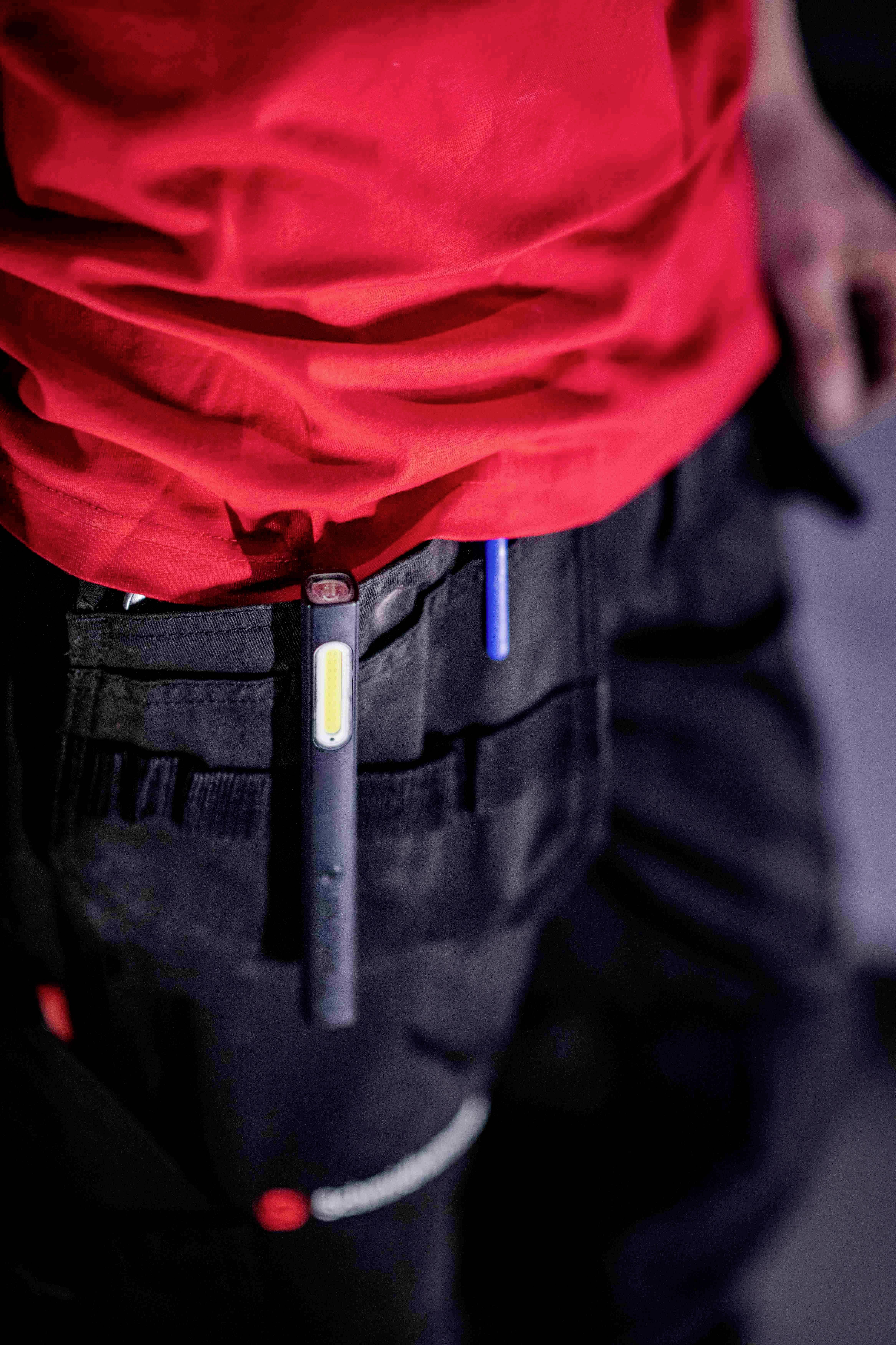 Une personne porte une veste de travail rouge et un pantalon noir avec des poches dans lesquelles sont glissés deux stylos.