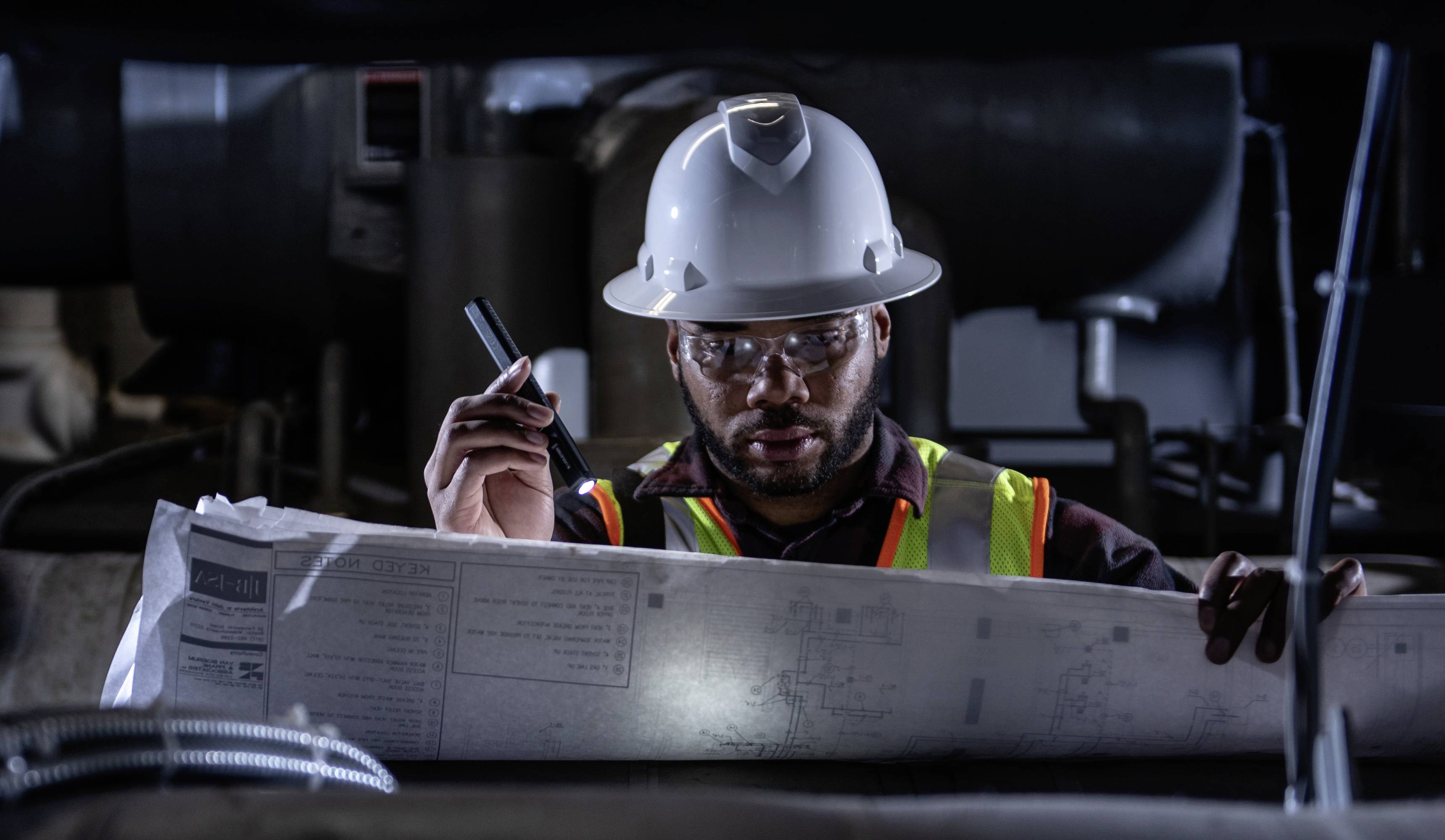 Un ouvrier porte un casque et un gilet de sécurité, tient une lampe de poche et étudie des plans de construction dans un environnement industriel.
