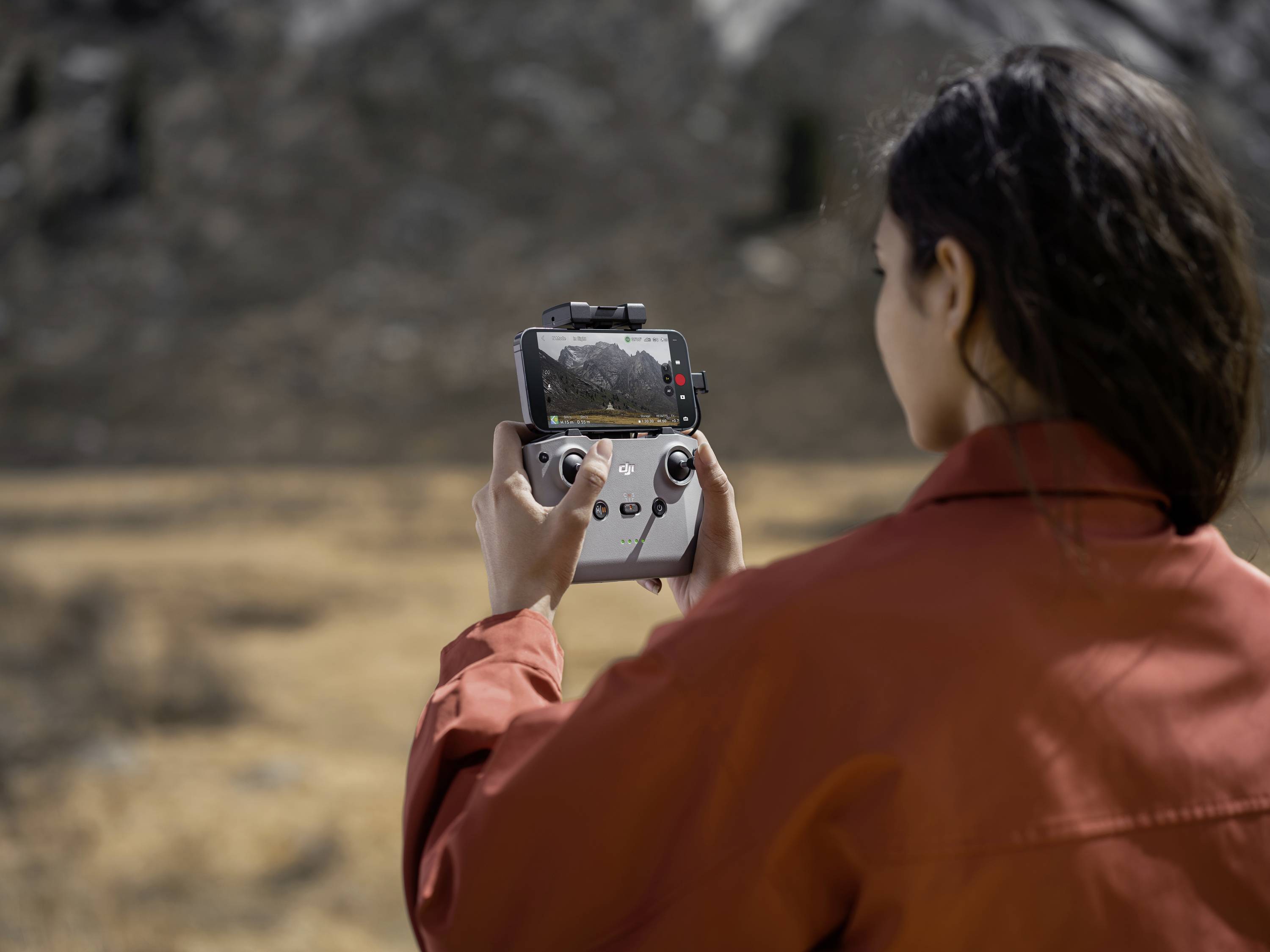 Une personne portant une veste rouge filme un panorama de paysage montagneux sur un écran à l'aide d'une télécommande de drone.