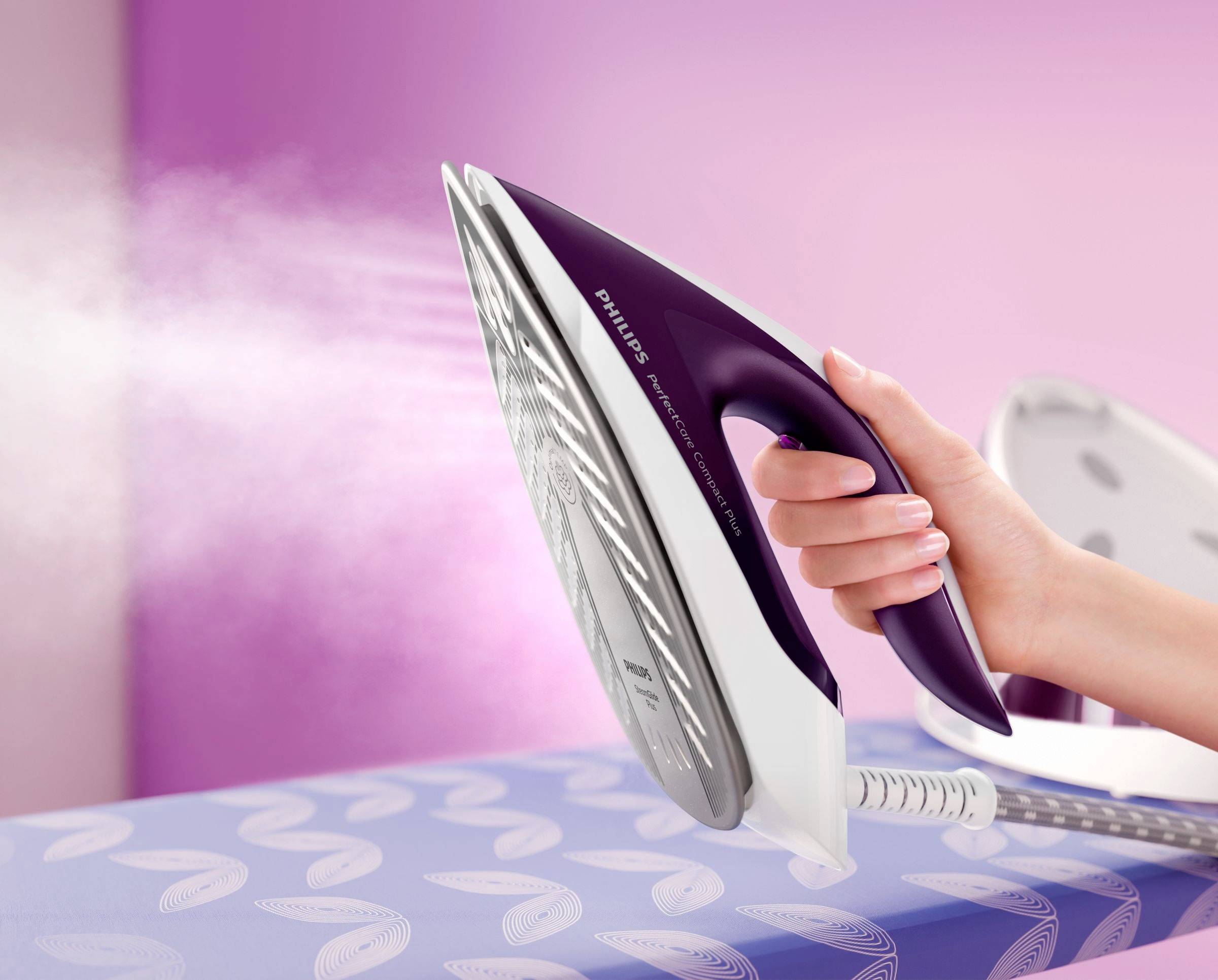 Une main tient un fer à repasser vapeur blanc et violet, qui dégage de la vapeur, au-dessus d'un tissu repassé sur une planche à repasser.