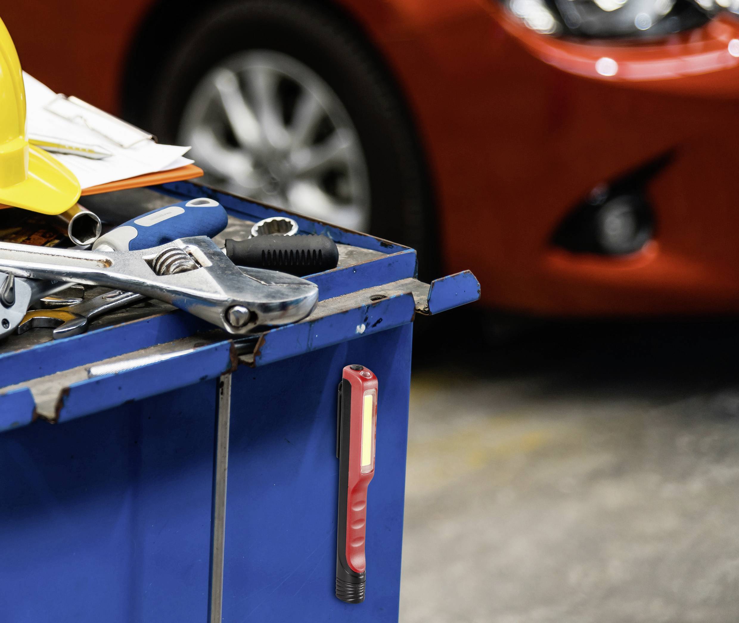 Un établi bleu avec des outils, dont des clés et un casque jaune. À l'arrière-plan, une voiture rouge dans un atelier.