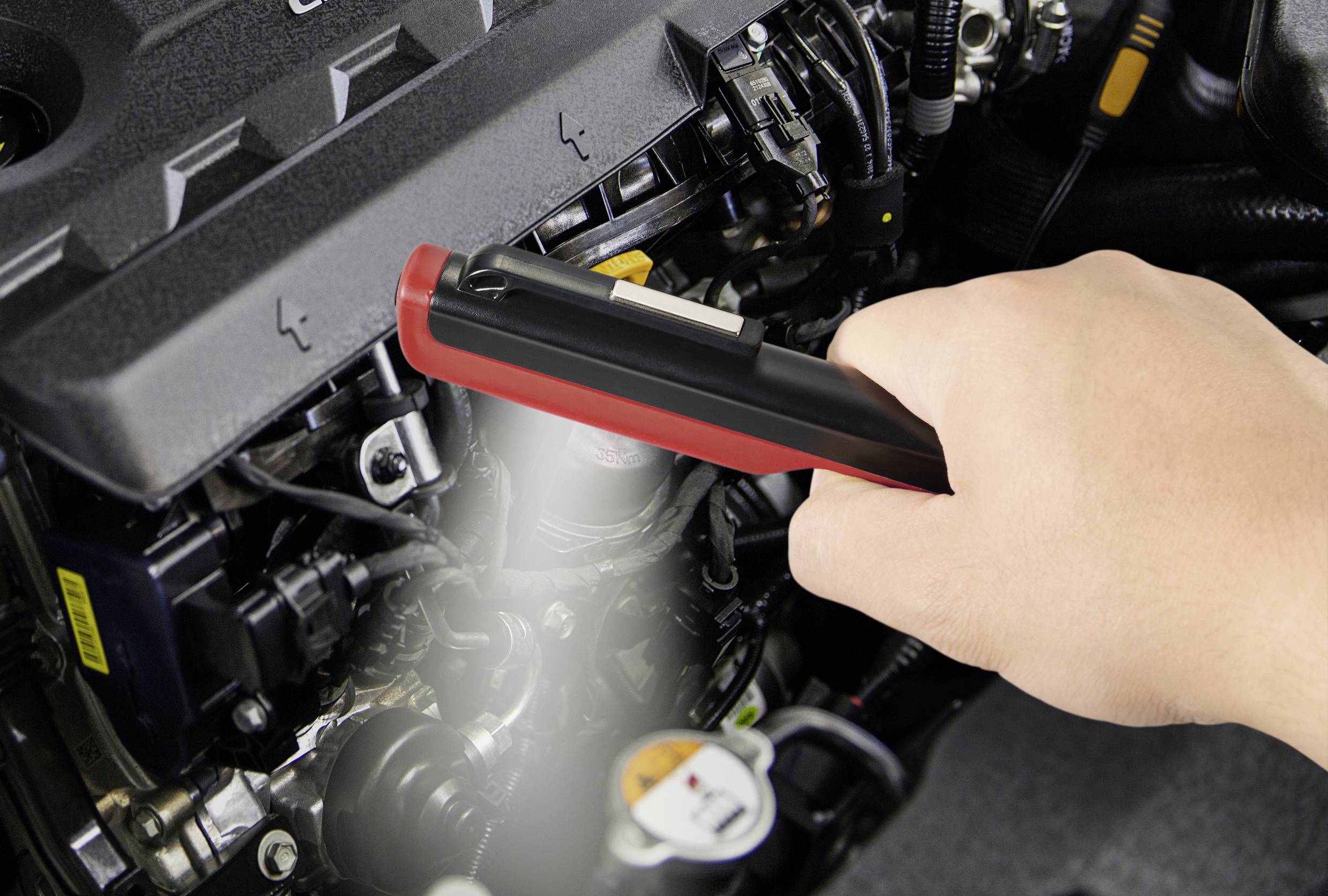 Une main utilise un outil de diagnostic pour vérifier le moteur d'une voiture. L'outil a un boîtier rouge et est tenu près du moteur.