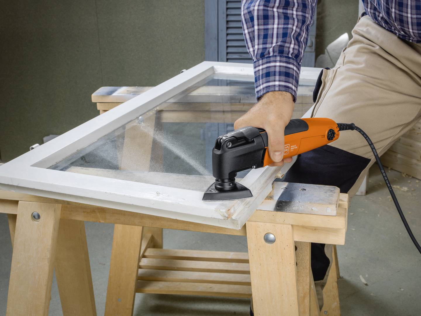 Une personne travaille un cadre de fenêtre avec un outil multifonction. Le cadre est posé sur un établi en bois à l'intérieur.