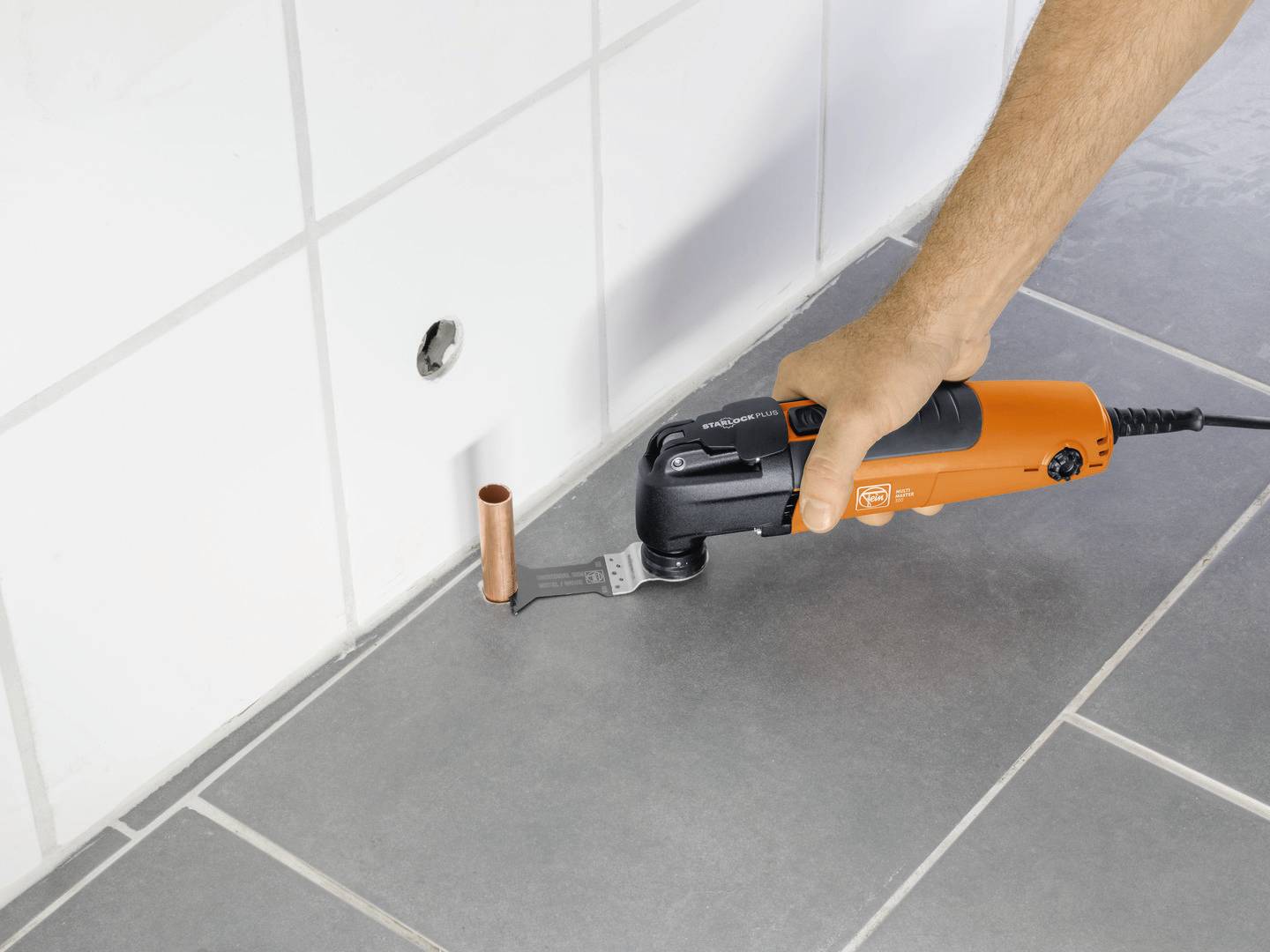 Une main tient un outil multifonction orange avec une lame près d'un tuyau en cuivre devant un mur carrelé.