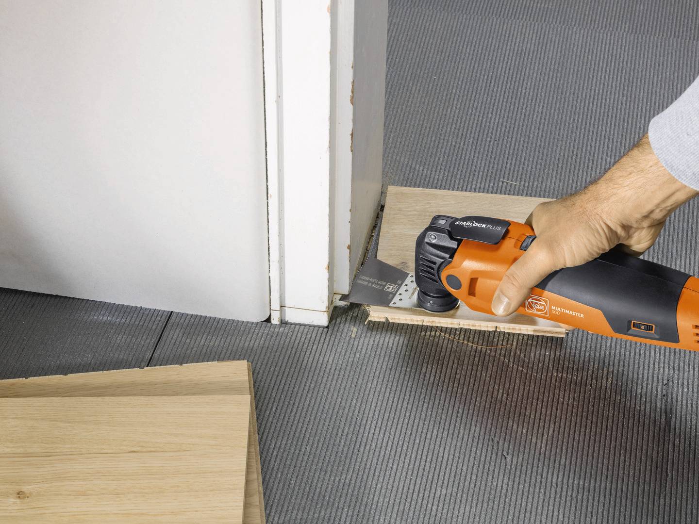 Une personne utilise un outil multifonction électrique pour couper un morceau de bois à côté d'une porte. Le sol est recouvert de carrelage gris.