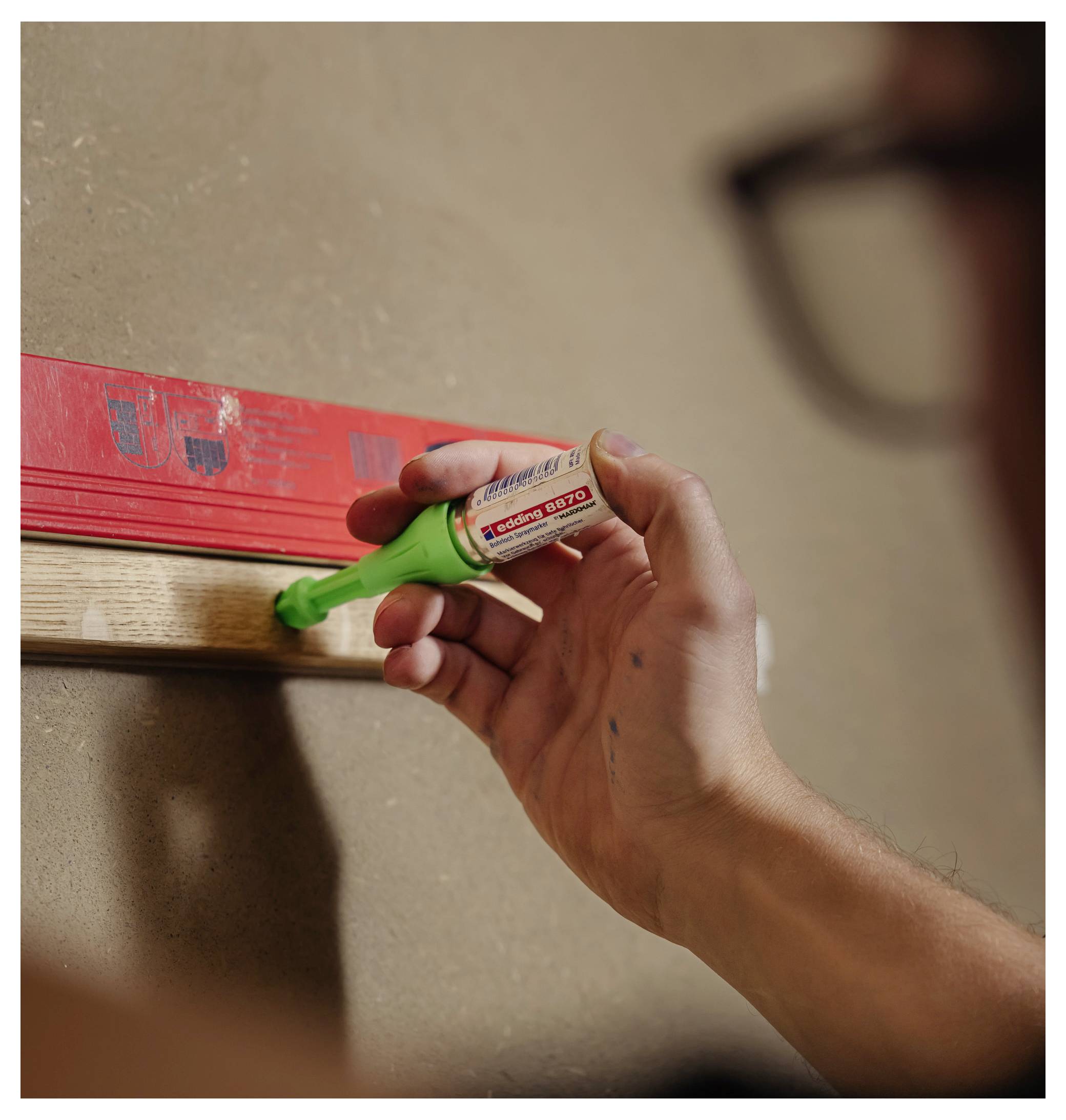 Une personne marquant une planche en bois avec un outil de marquage vert pour charpentier tout en mesurant contre un niveau rouge.