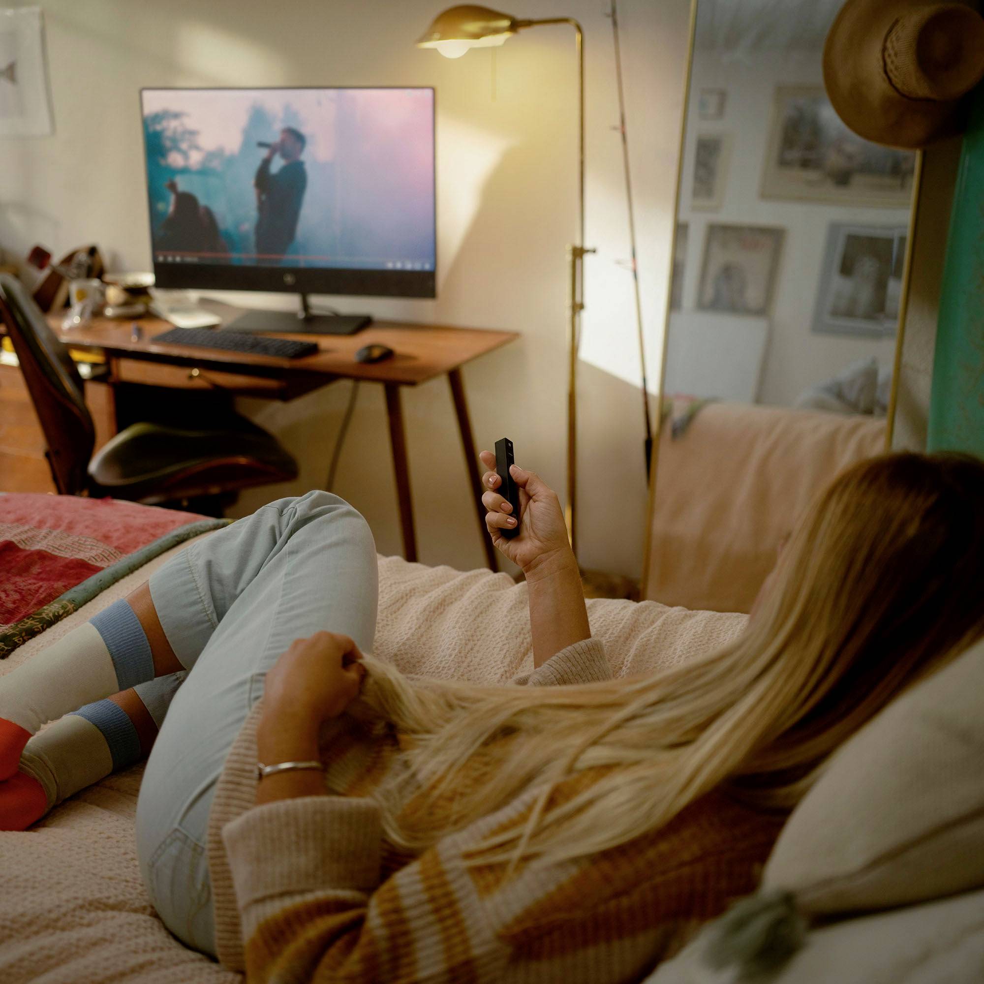 Une femme est allongée sur un lit et regarde la télévision. Un bureau avec un écran est visible en arrière-plan. L'atmosphère de la chambre est confortable et détendue.