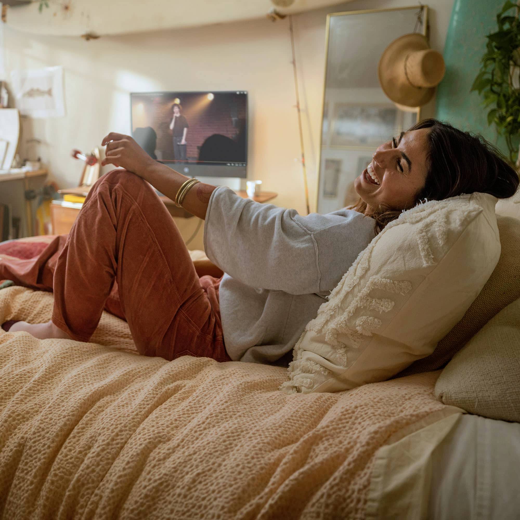 Une personne souriante est allongée de manière détendue sur un lit, regardant quelque chose à la télévision. La pièce semble chaleureuse et accueillante.