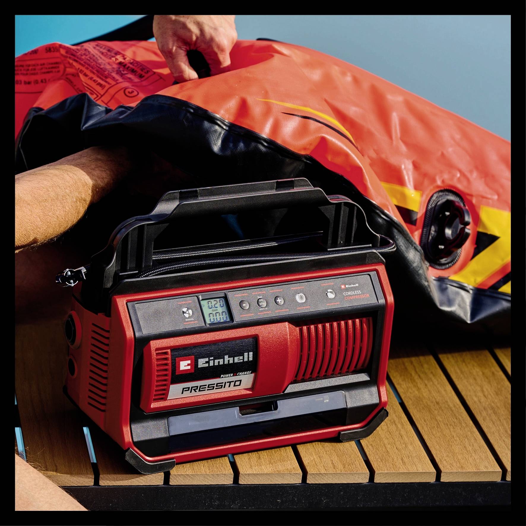 Une pompe à air portable rouge avec affichage numérique et boutons de commande est utilisée pour gonfler un matelas de piscine orange sur une terrasse en bois.