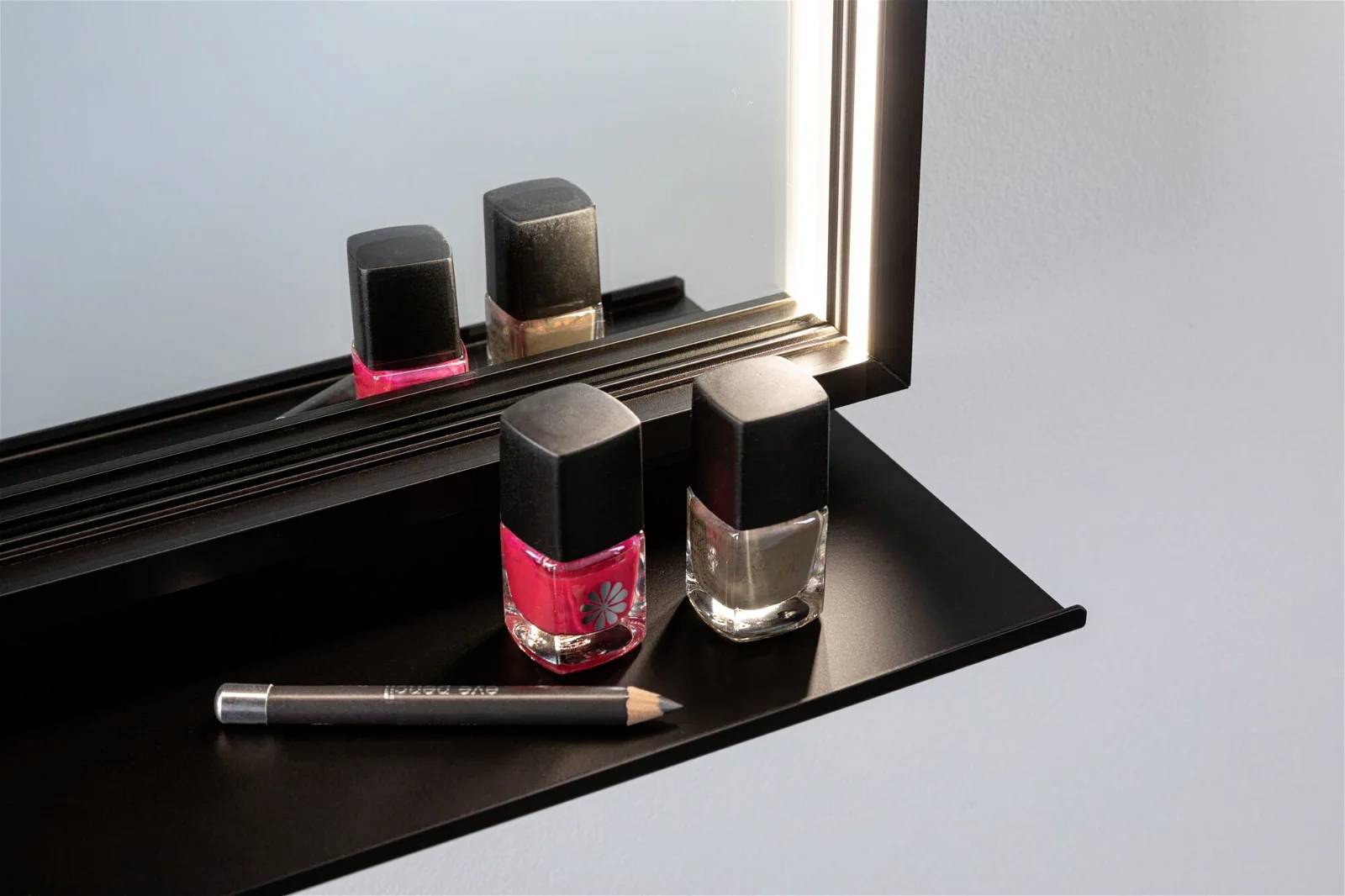 Deux petits flacons de vernis à ongles, l'un rouge et l'autre transparent, sont posés sur une étagère noire devant un miroir. Un stylo noir est placé à côté.