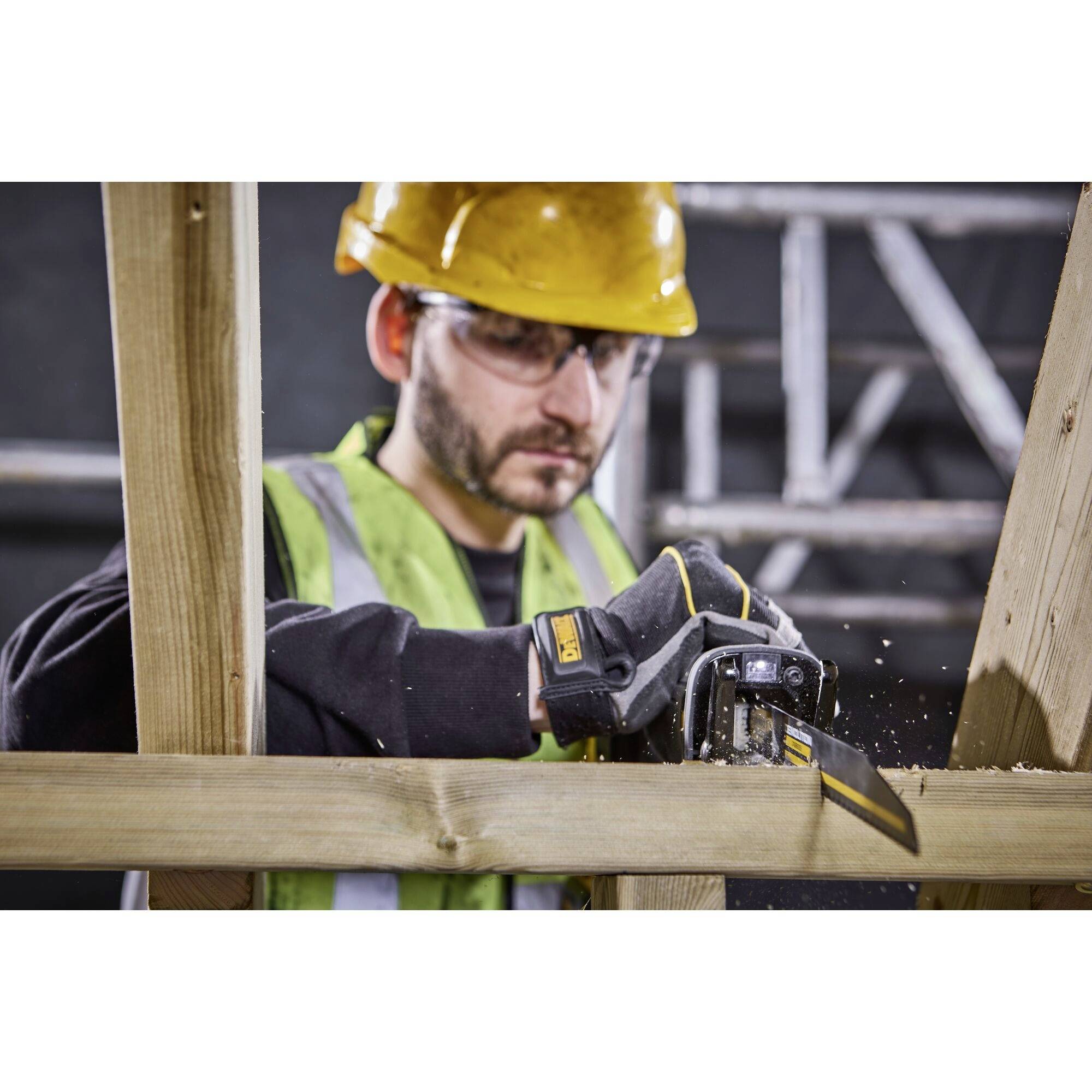 Un ouvrier en tenue de protection, portant un casque et des lunettes, utilise une scie à main pour couper du bois. Un échafaudage est visible en arrière-plan.