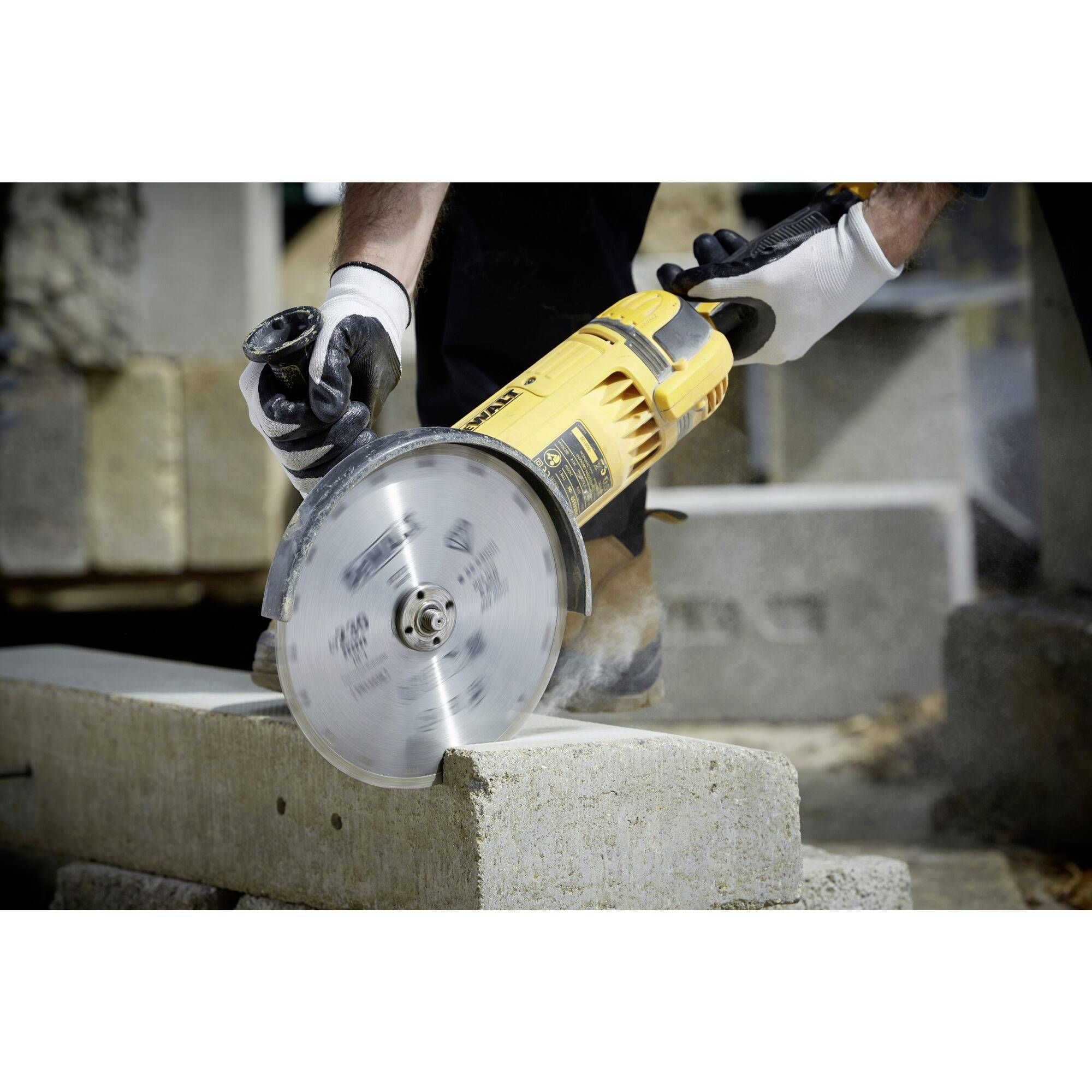 Une personne coupe un bloc de béton avec un meuleuse d'angle électrique, porte des gants de protection et travaille à l'extérieur.