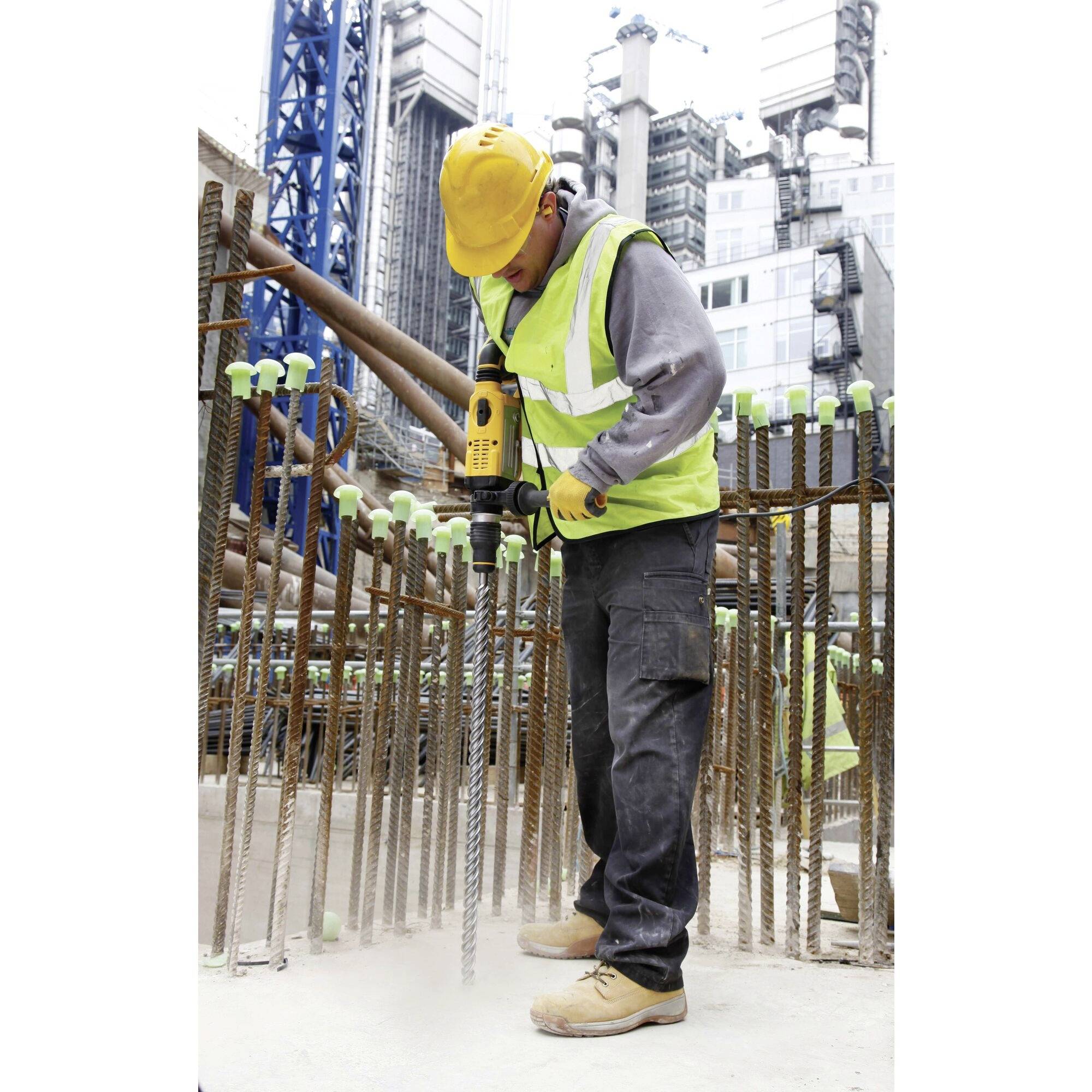 Un ouvrier en tenue de protection fore des trous dans du béton avec un marteau-piqueur sur un grand chantier de construction, avec des poutres d'acier en arrière-plan.