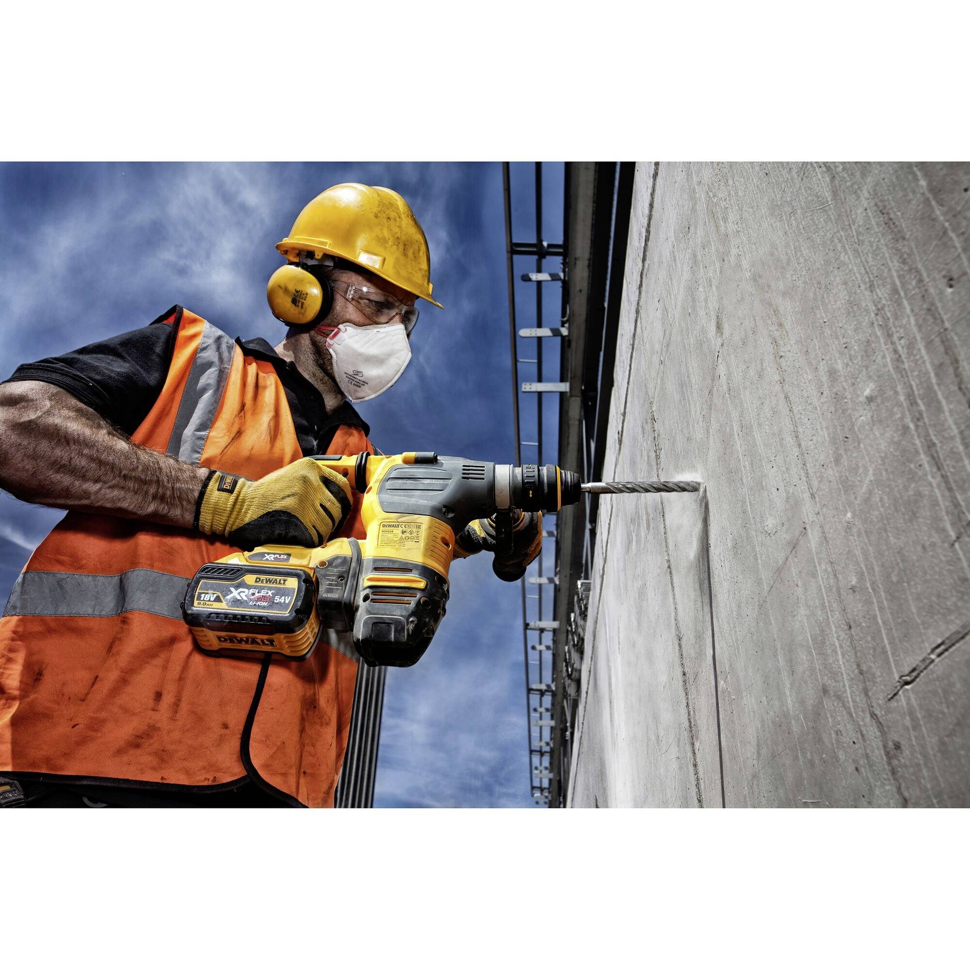 Un ouvrier portant un casque et des lunettes de protection fore un mur en béton avec une grande perceuse. Un ciel bleu se trouve en arrière-plan.