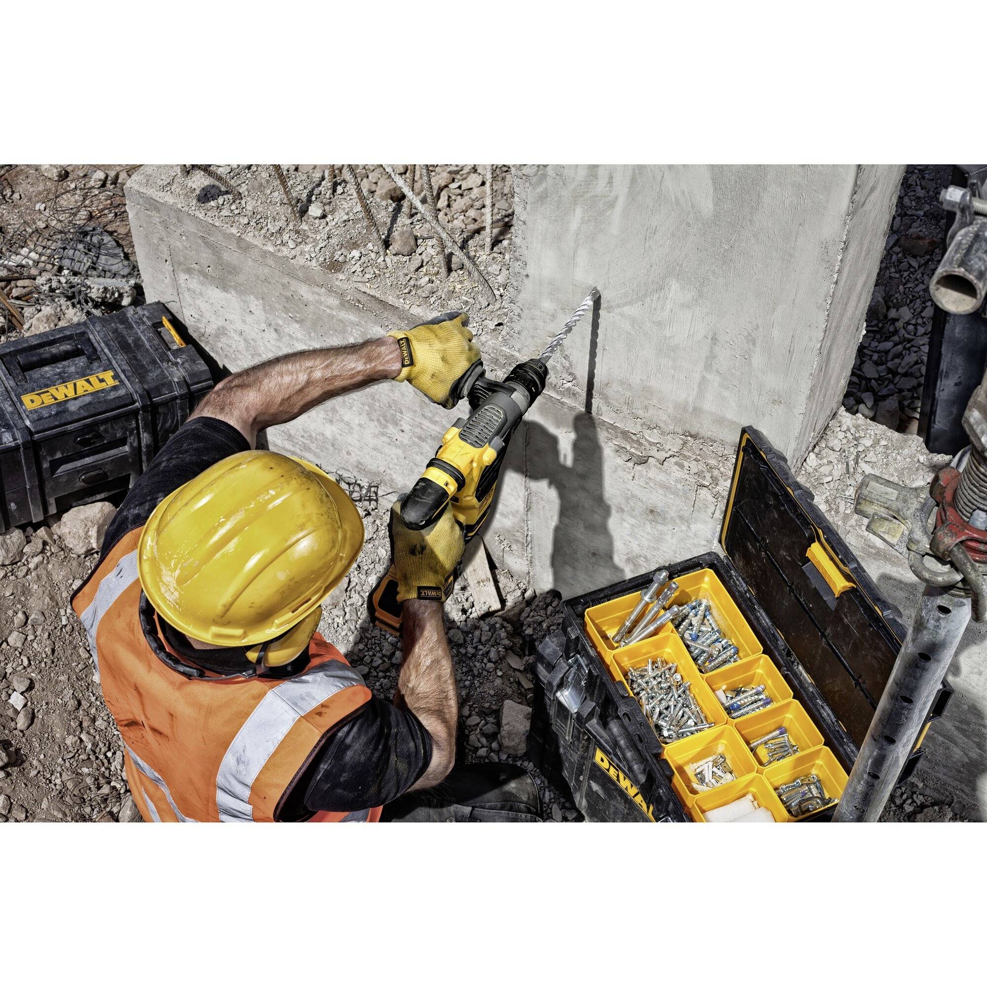 Une personne vêtue de vêtements de chantier et portant un casque utilise une perceuse pour percer du béton. À côté d'elle se trouve une boîte à outils contenant des vis.
