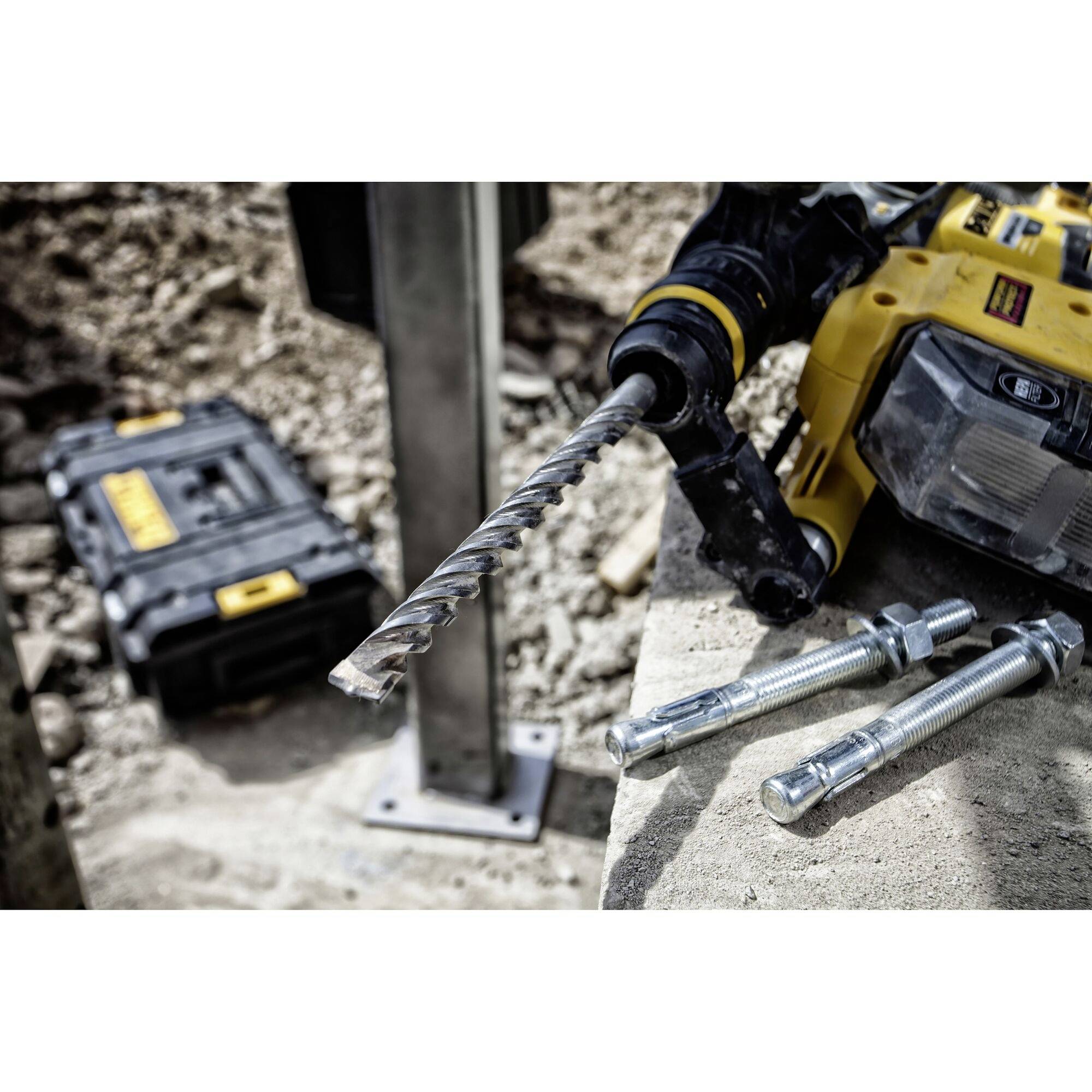 Un marteau-piqueur jaune est posé sur un chantier, à côté de deux boulons d'ancrage et d'une boîte à outils en arrière-plan.