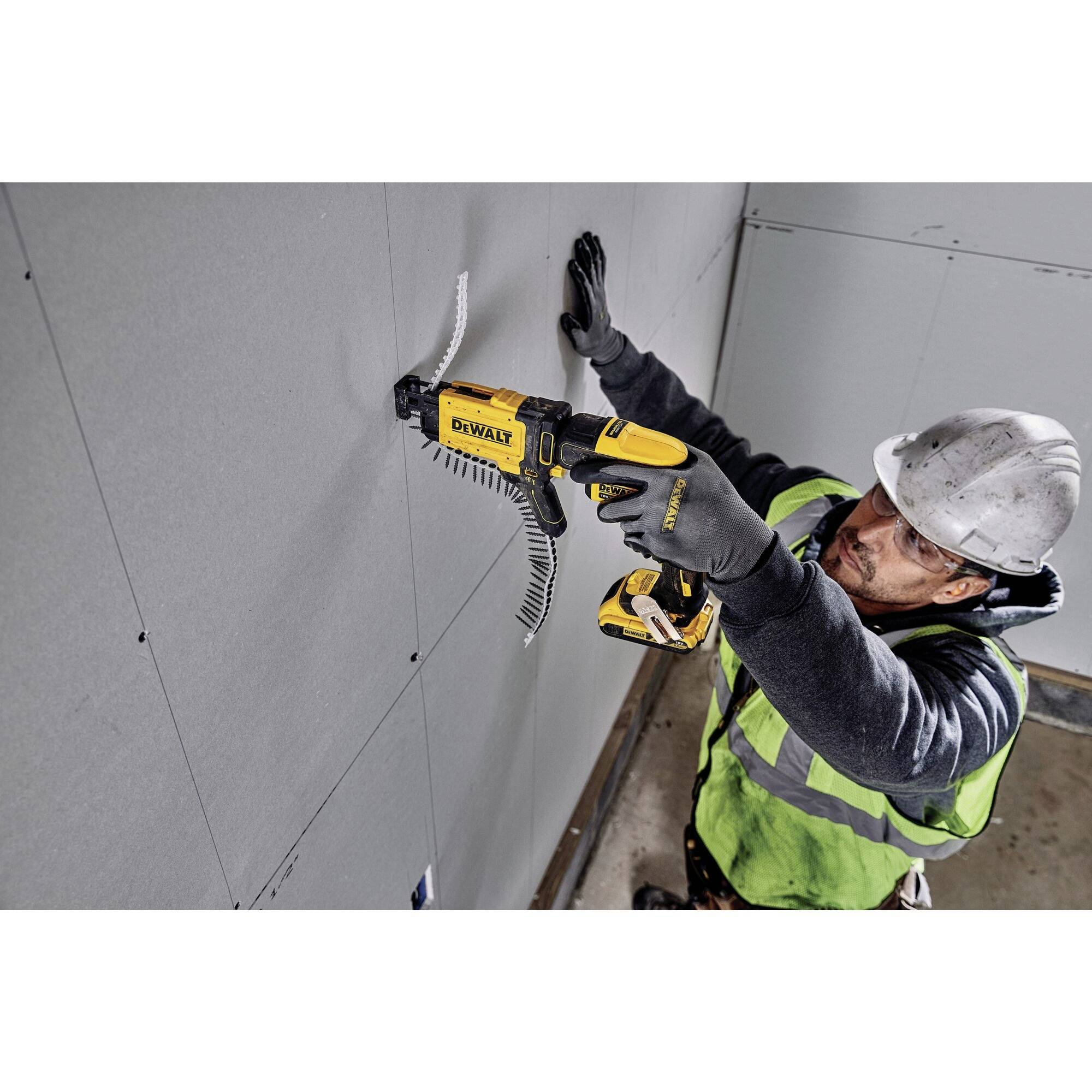 Un ouvrier portant un casque et un gilet de protection visse une cloison en plaque de plâtre. Il utilise un outil électrique jaune et porte des gants.