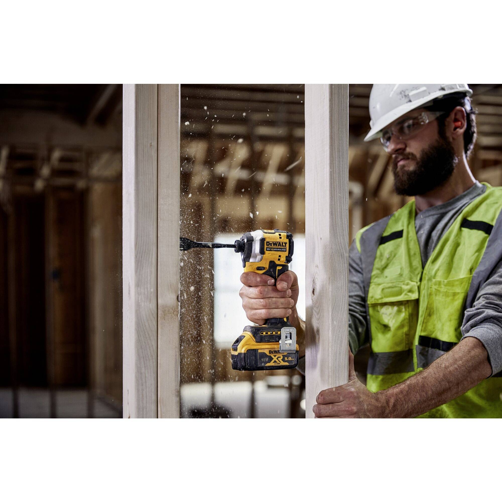 Un ouvrier portant un casque de chantier et un gilet de sécurité fore une planche en bois dans un bâtiment en construction. Il tient une perceuse jaune à la main.