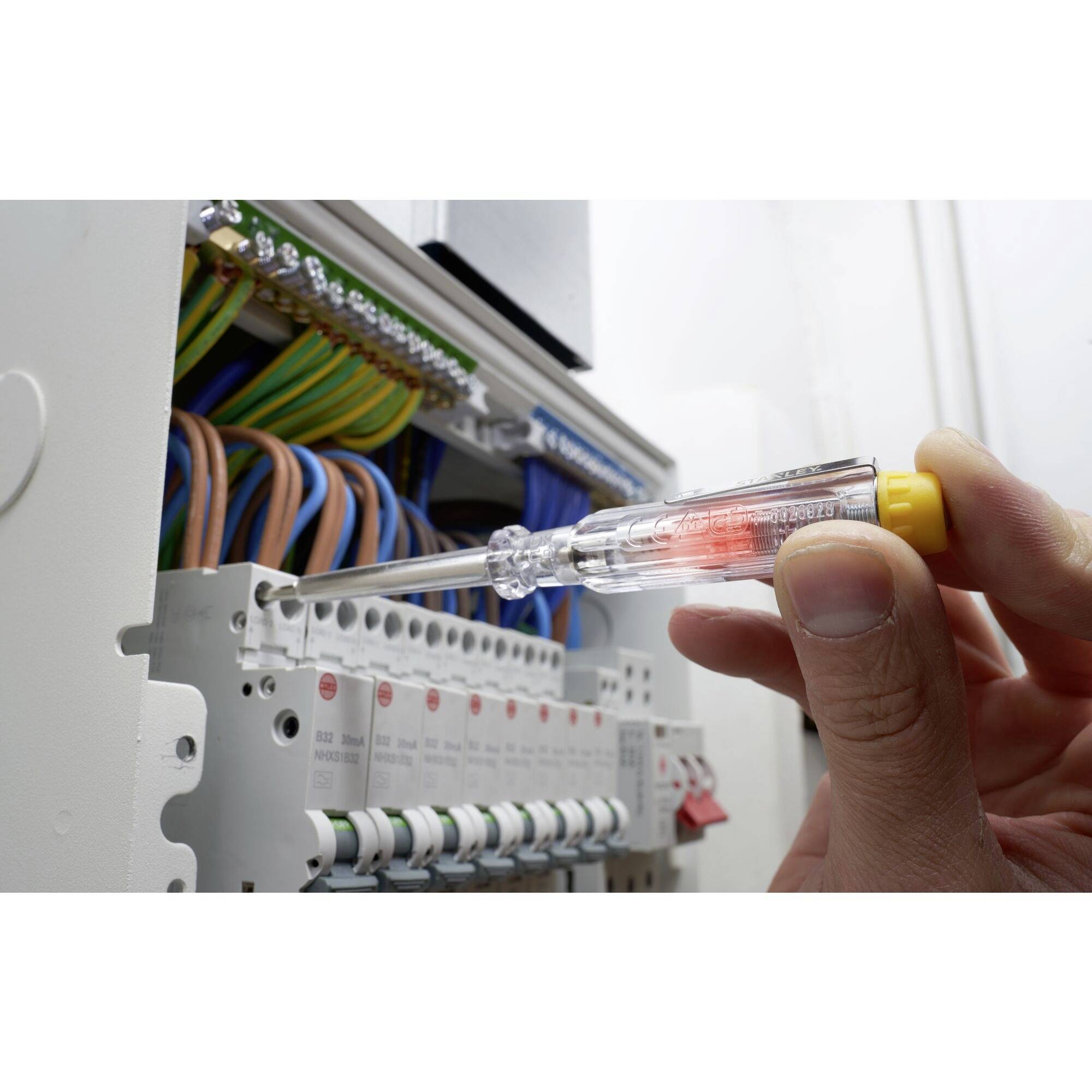 Une main tient un testeur de phase qui s'illumine en rouge, près d'un distributeur électrique ouvert avec des câbles de couleurs pour vérifier la tension.