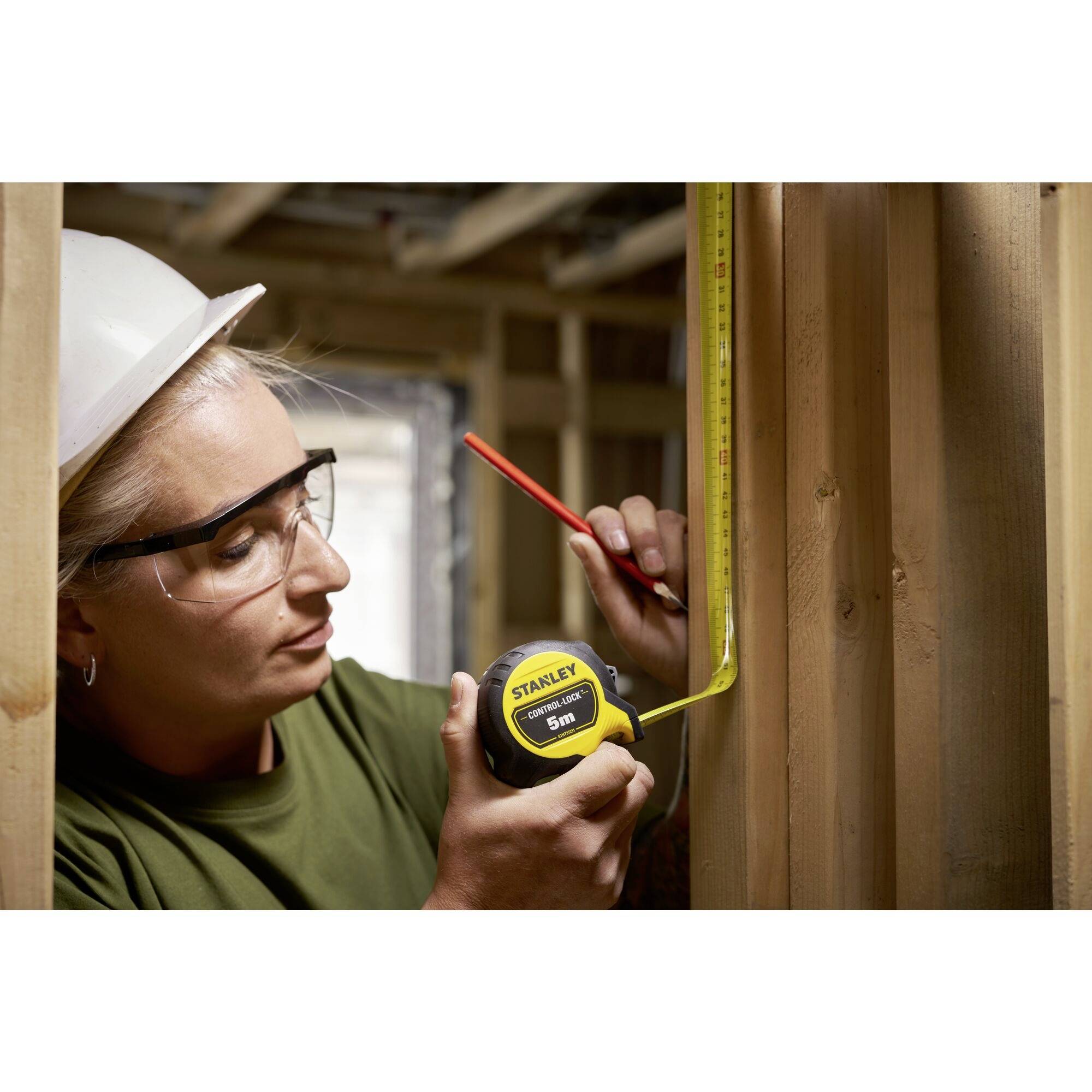 Une personne portant un casque de protection et des lunettes mesure avec un mètre ruban et marque avec un stylo des cadres en bois dans une pièce en cours de finition.