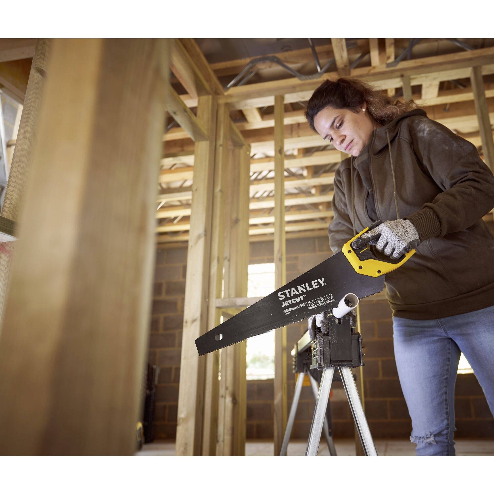 Une femme en tenue de travail utilise une scie à main sur un chantier. Elle se tient devant un cadre en bois et est concentrée dans son travail.