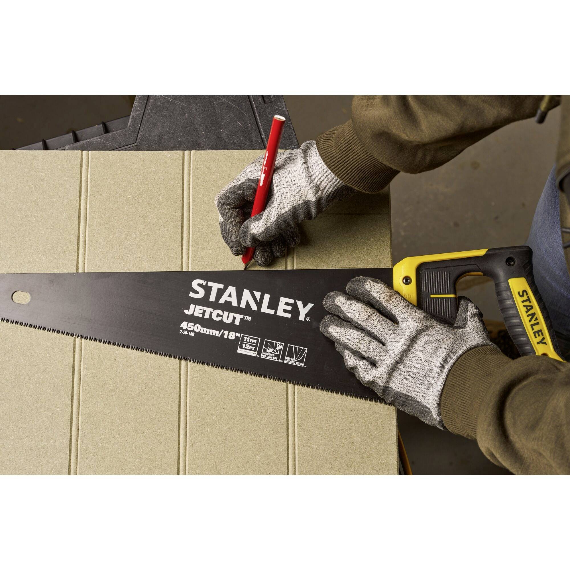 Une personne portant des gants gris dessine avec un stylo rouge sur du bois, à côté d'une scie à main noire de la marque Stanley.