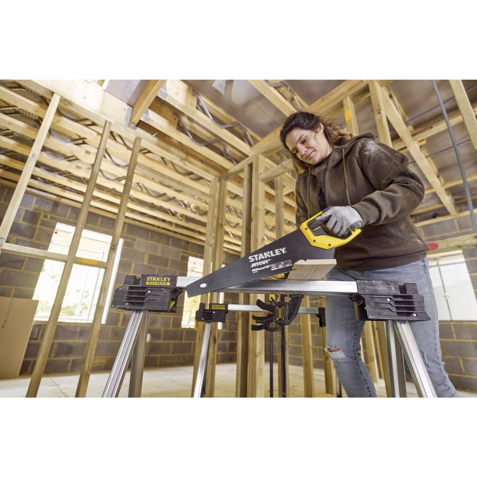 Une personne scie un morceau de bois dans un bâtiment en construction à l'aide d'une scie à main. Des cadres en bois et des murs inachevés sont visibles en arrière-plan.