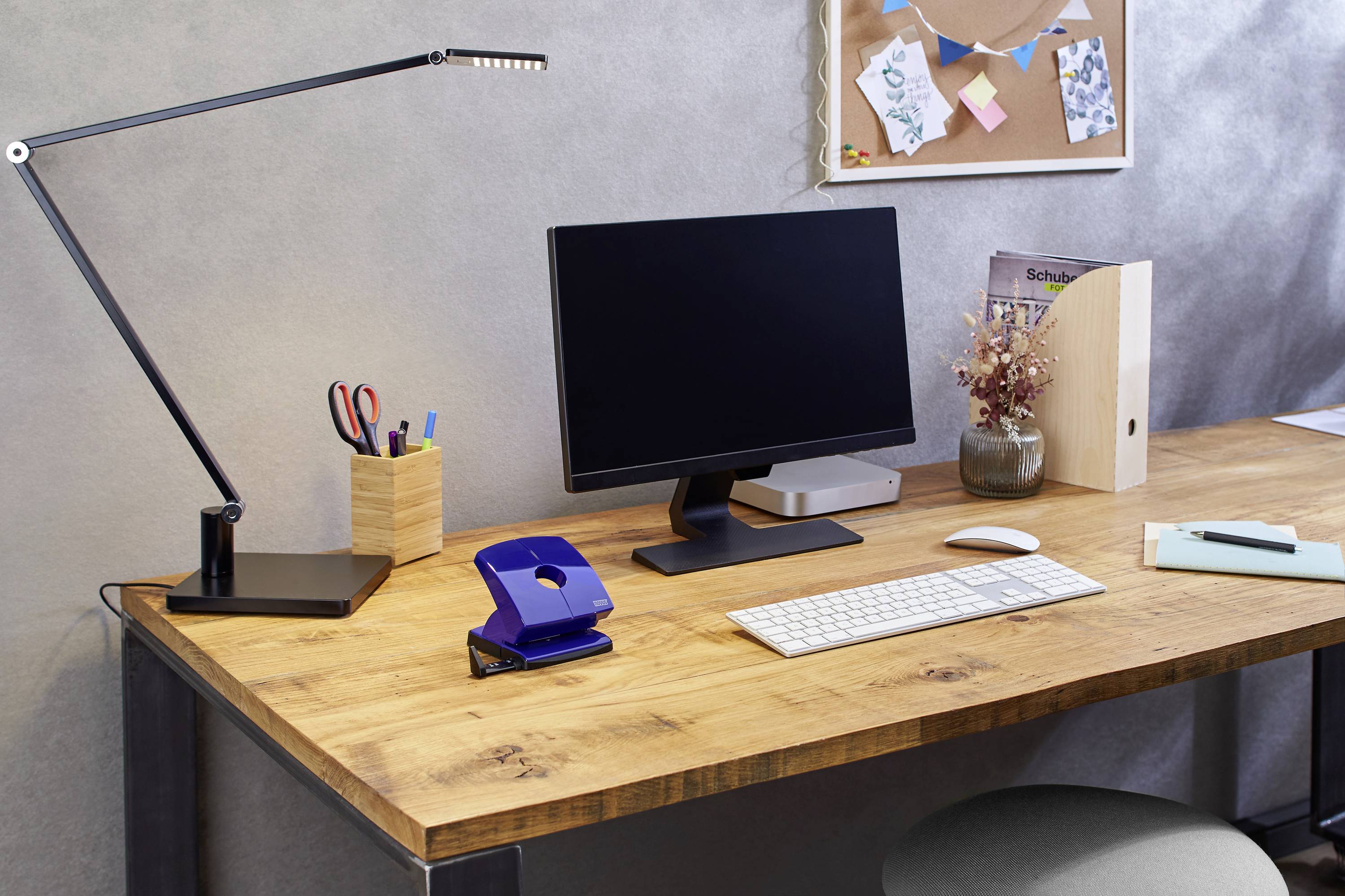 Bureau avec lampe, écran, clavier, souris, porte-stylos, agrafeuse et éléments décoratifs. En arrière-plan, un tableau d'affichage.