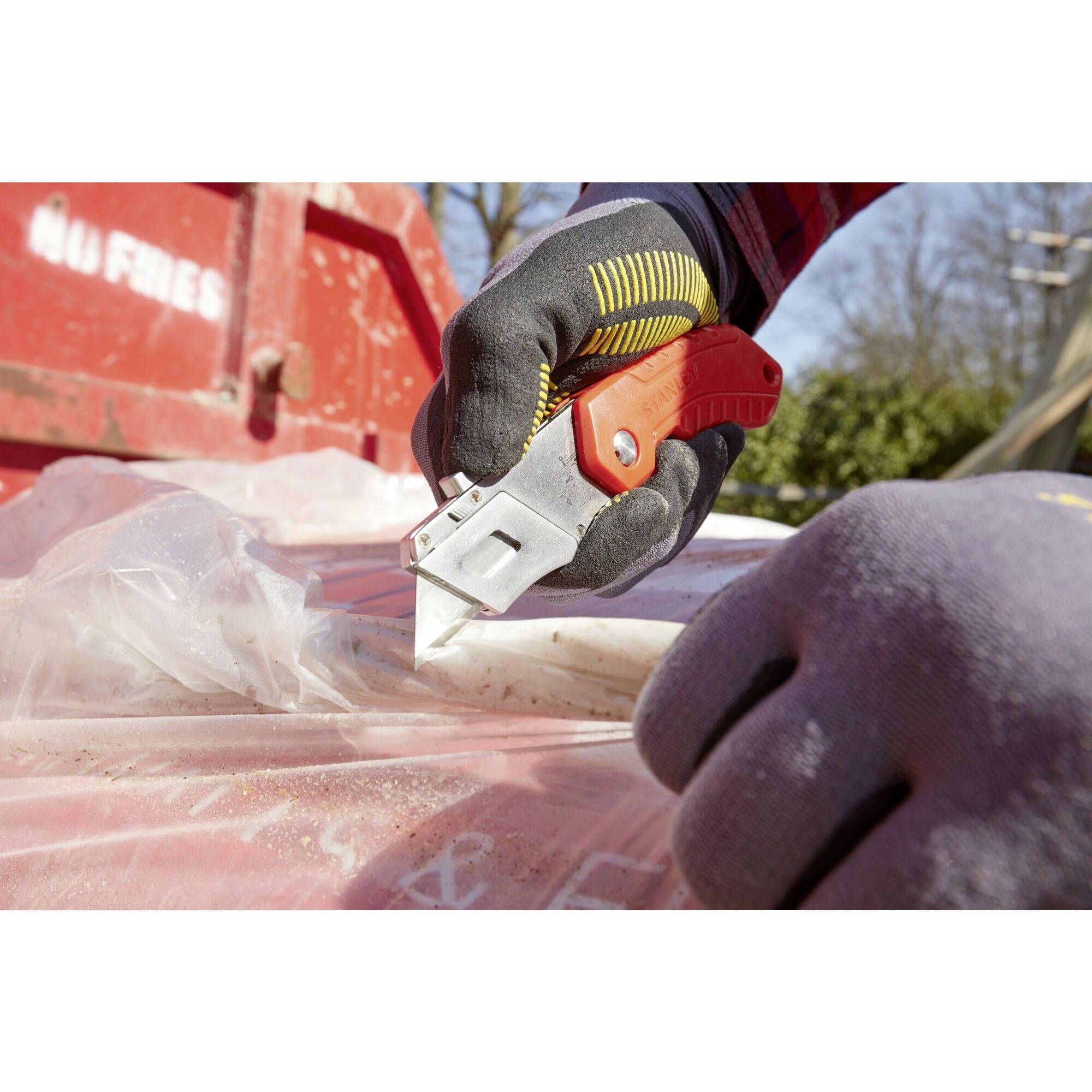 Une personne coupe du plastique avec un cutter sur un conteneur rouge. Des vêtements de protection et des gants sont portés.