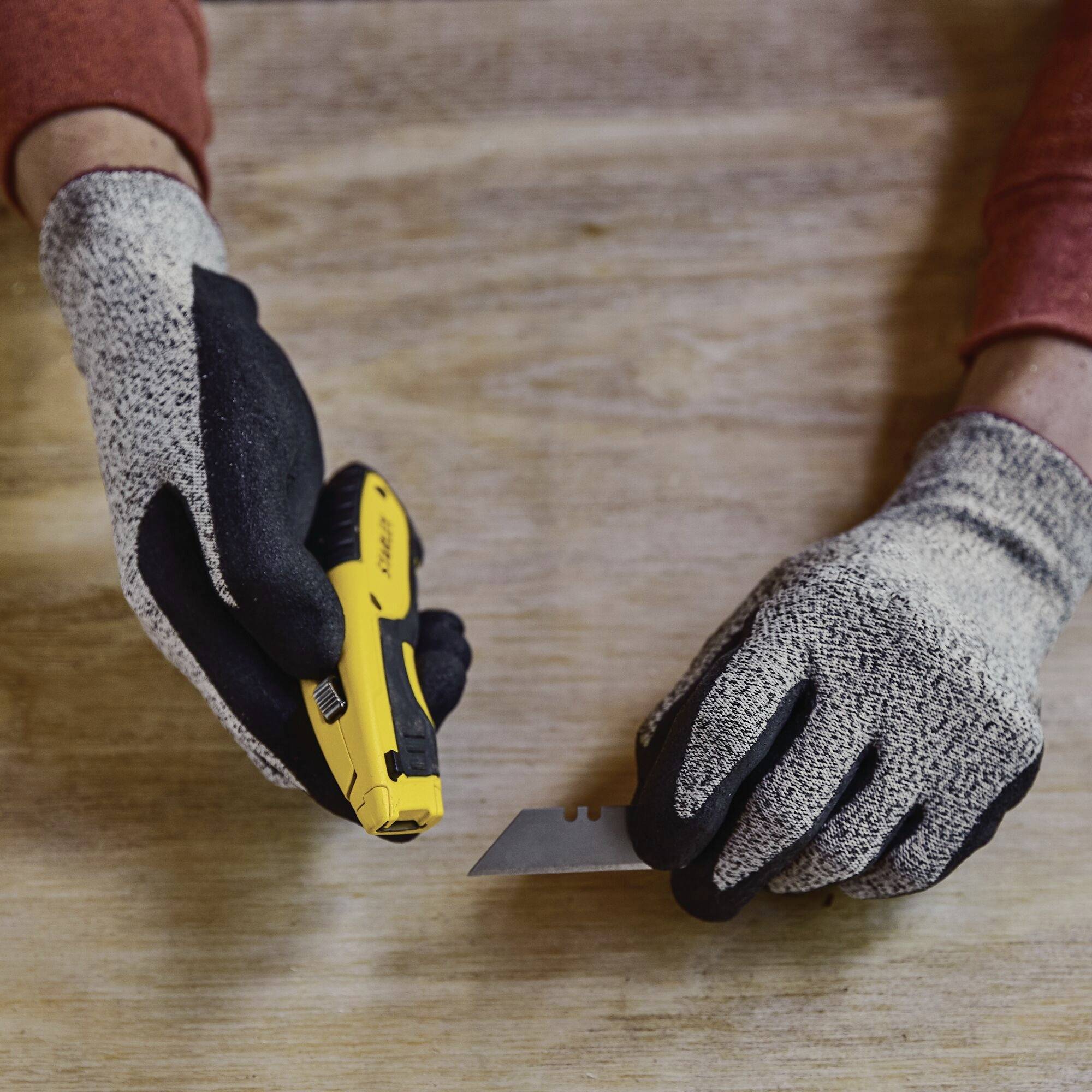 Deux mains gantées tiennent un cutter jaune glissant sur une surface en bois ; focus sur un travail de précision.