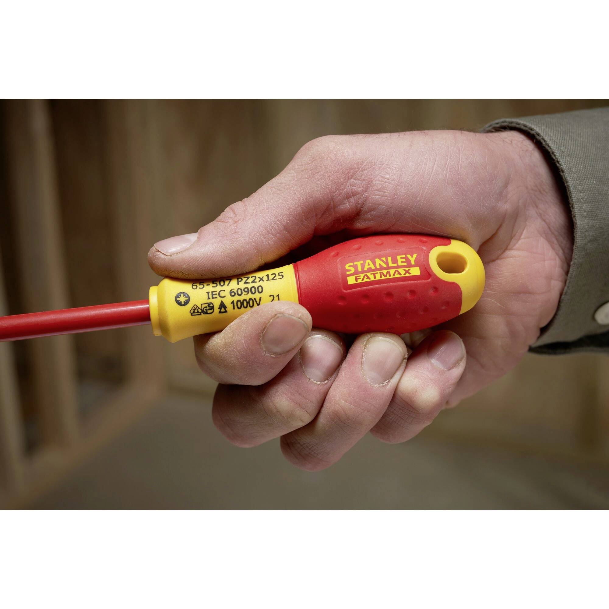 Une main tient un tournevis jaune et rouge de la marque 'Stanley Fatmax', adapté pour des tensions jusqu'à 1000V, dans une pièce aux murs en bois.