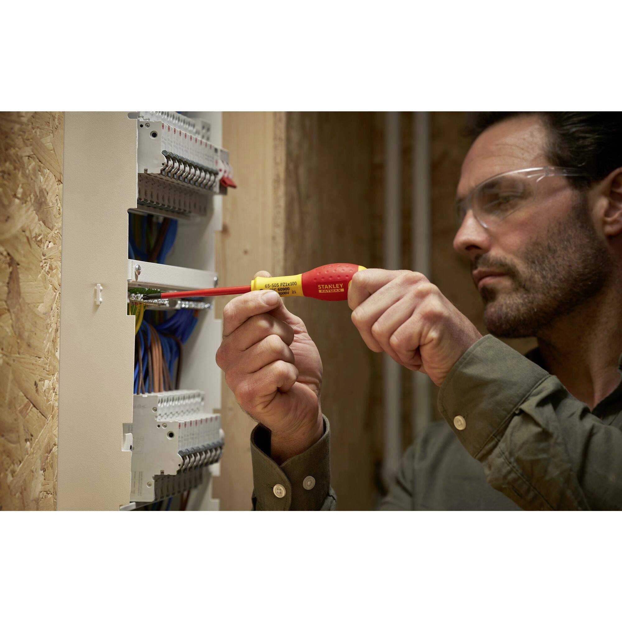 Un électricien travaille avec un tournevis sur un tableau électrique ouvert et porte des lunettes de protection.